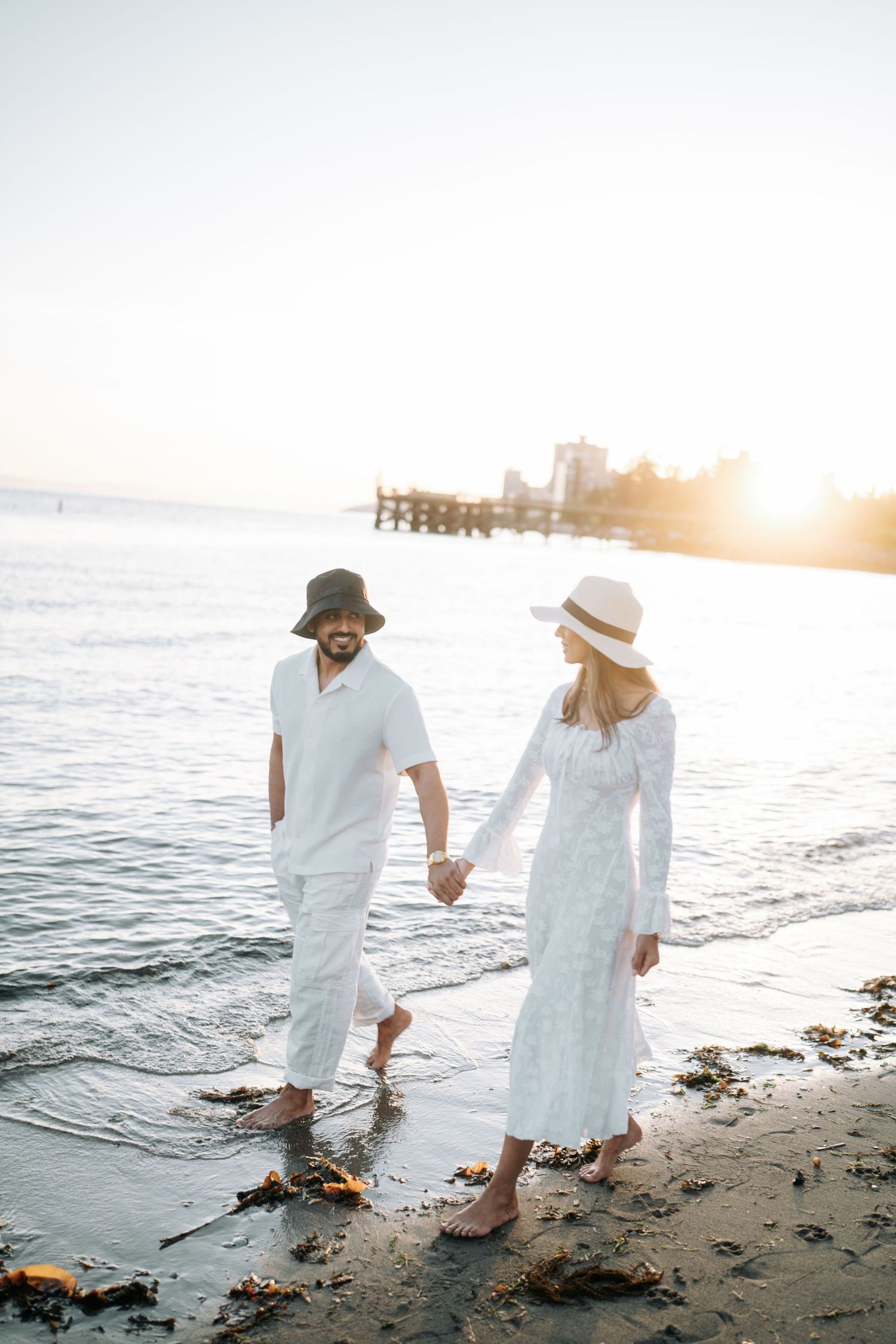 Couple in white outfits walking along a beach shoreline during sunset creating a romantic coastal atmosphere.