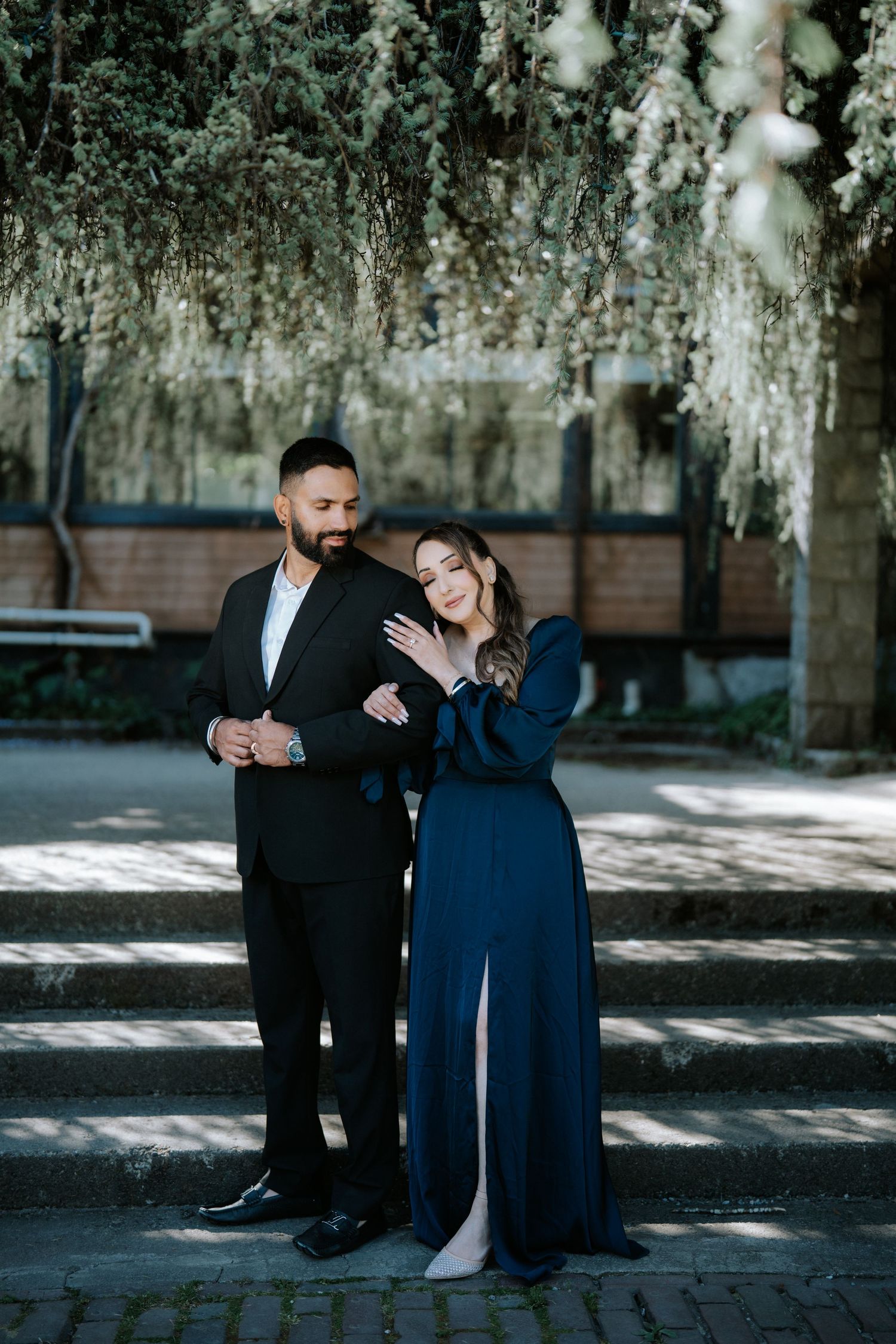 Elegant couple posing outdoors under hanging white flowers, man in black suit, woman in navy blue gown.