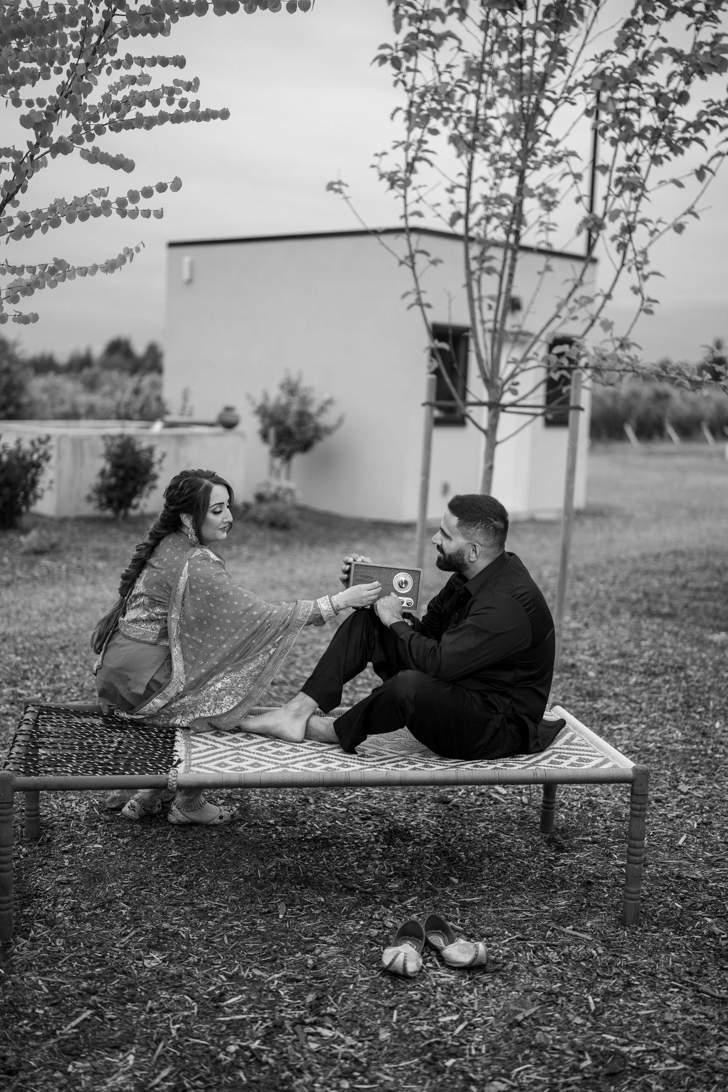 Black and white photo of a couple in traditional South Asian attire sitting on a charpai outdoors, sharing a warm moment.