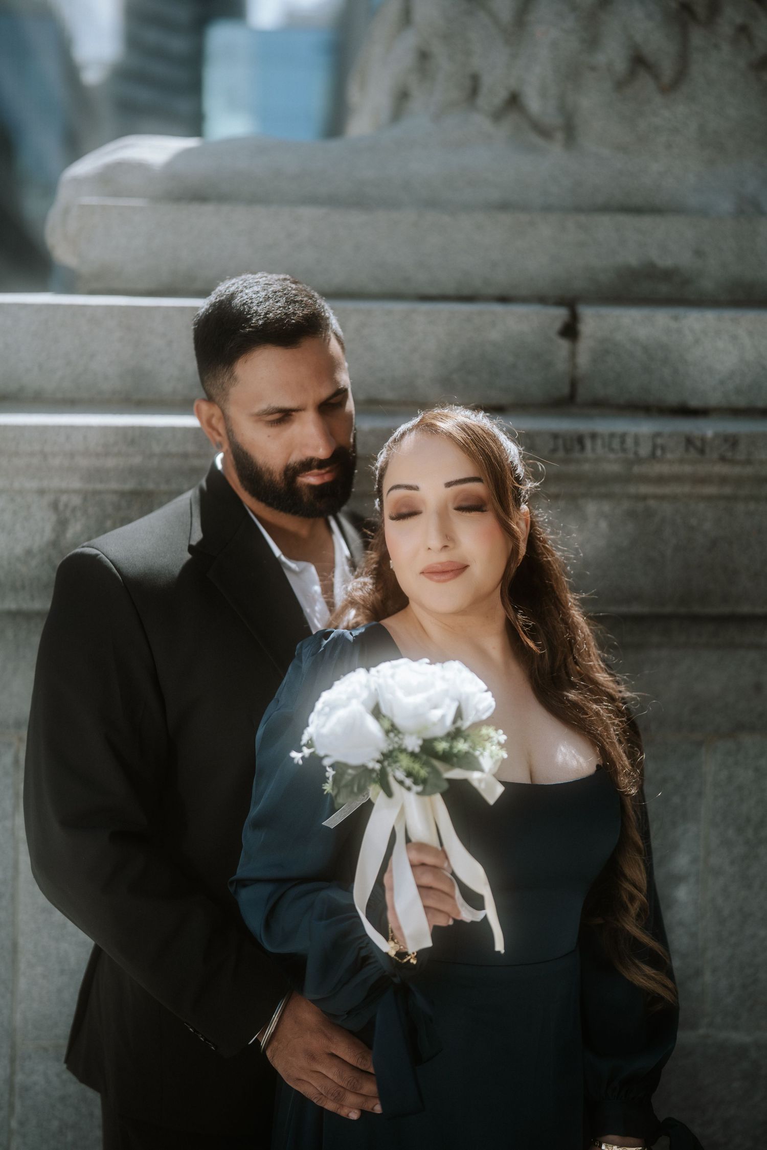 Couple in elegant attire poses romantically outdoors, woman in black dress holding white rose bouquet, man in black suit beside her.