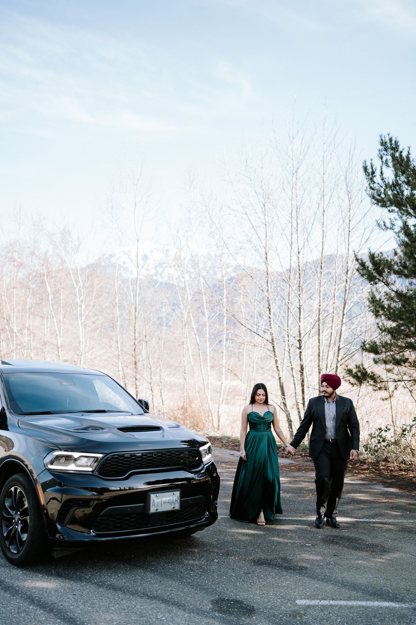 A couple poses near a black car on a winter day with bare trees in the background.