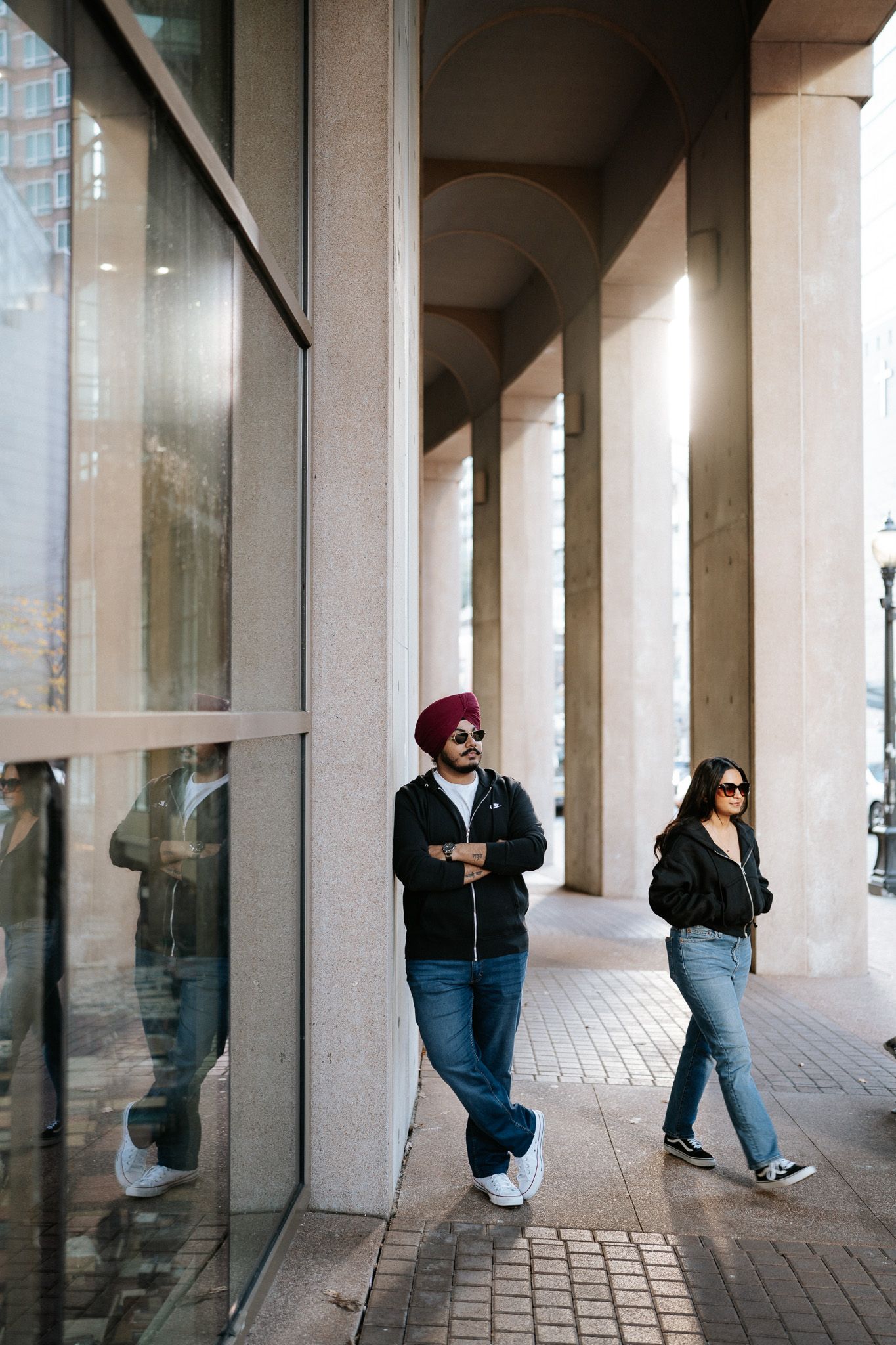 People stand in a covered walkway with white columns and glass windows creating a repeating architectural pattern.