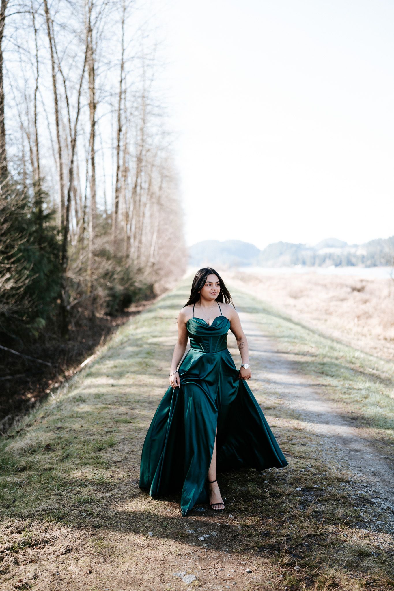 A person in an elegant teal evening gown poses on a rustic dirt path lined with bare winter trees.