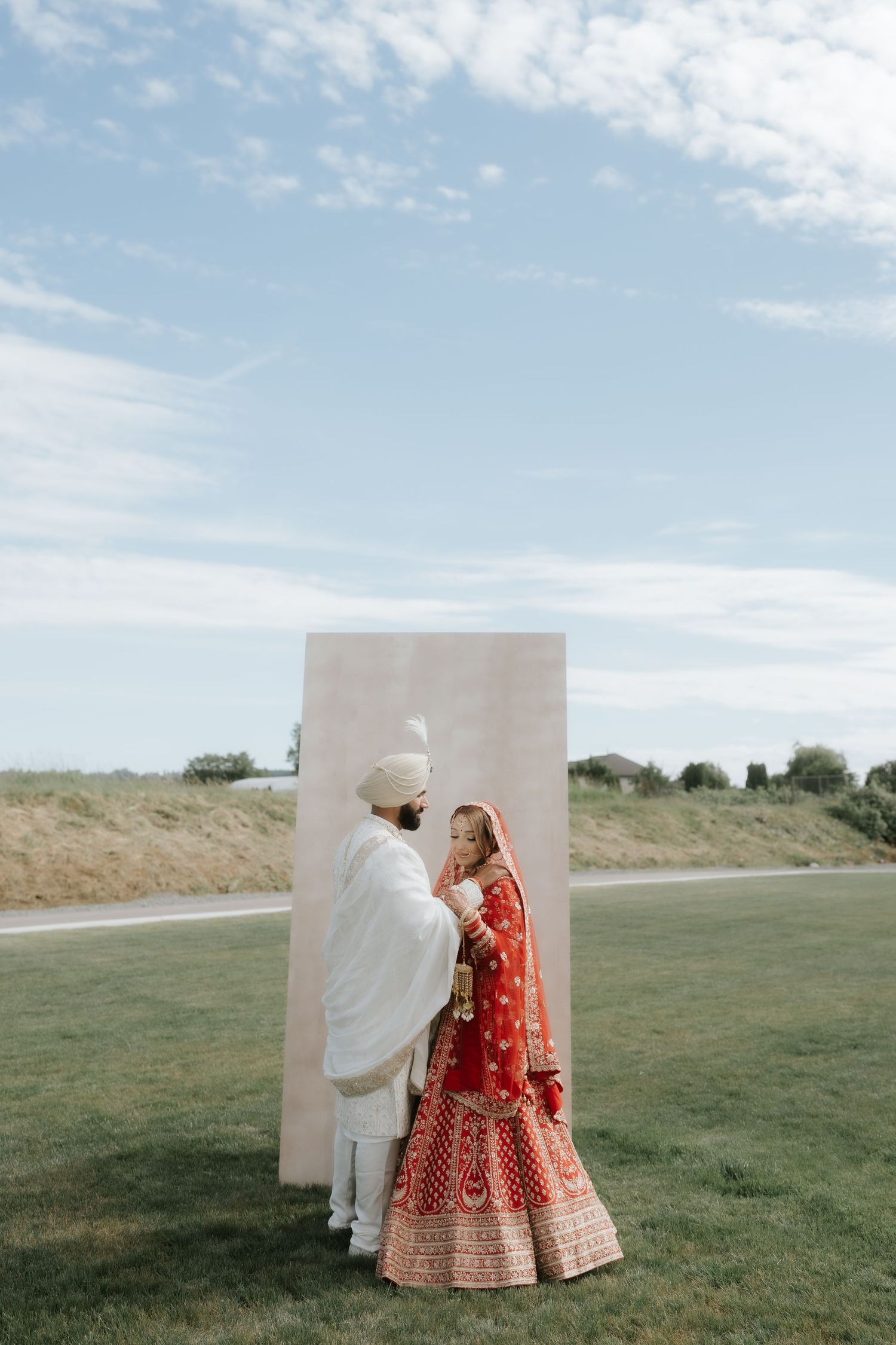 Indian bride in red lehenga and groom in white sherwani share a first look moment outdoors on a sunny day.