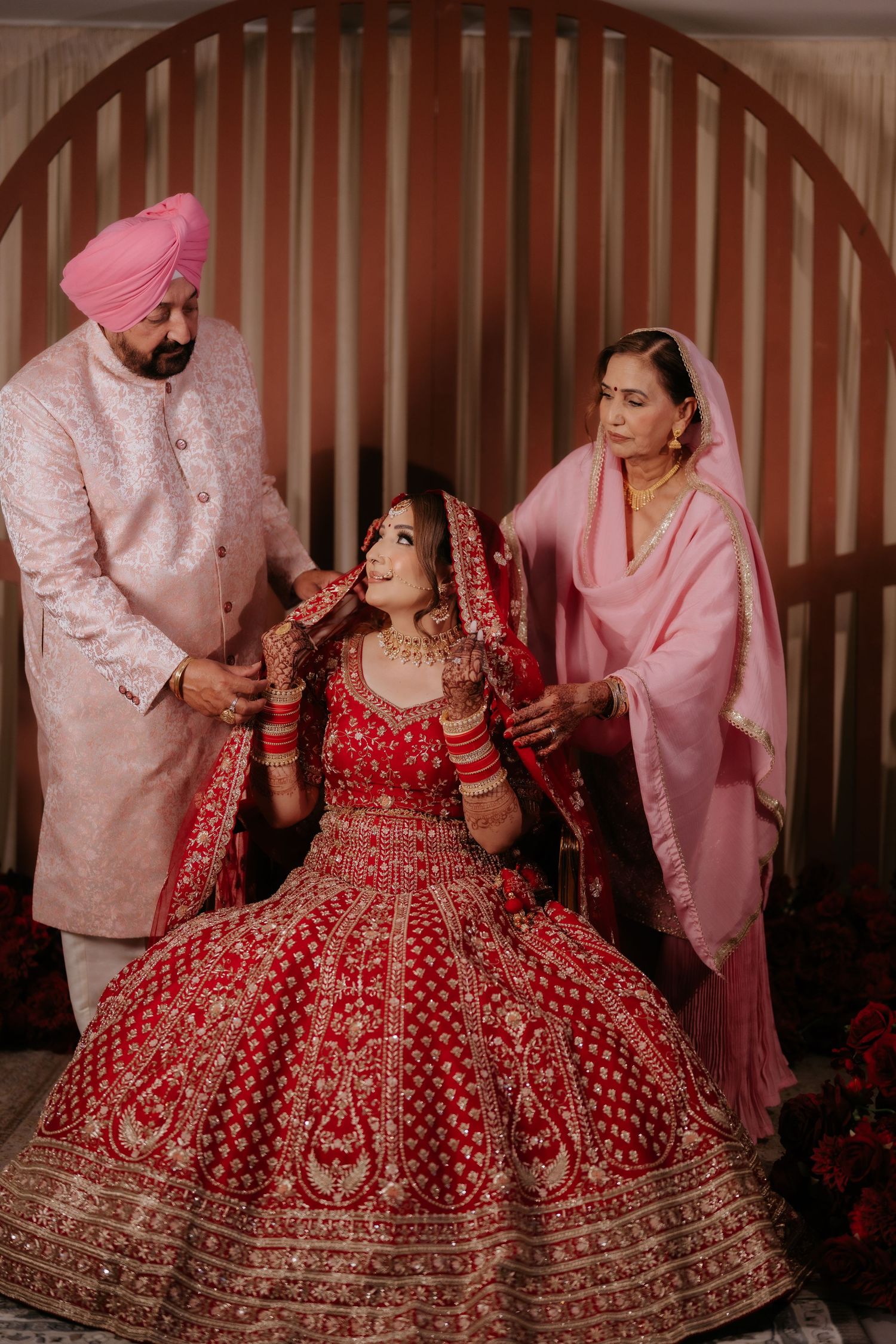 Indian bride in red lehenga seated while parents assist with bridal jewelry during traditional Punjabi wedding ceremony.