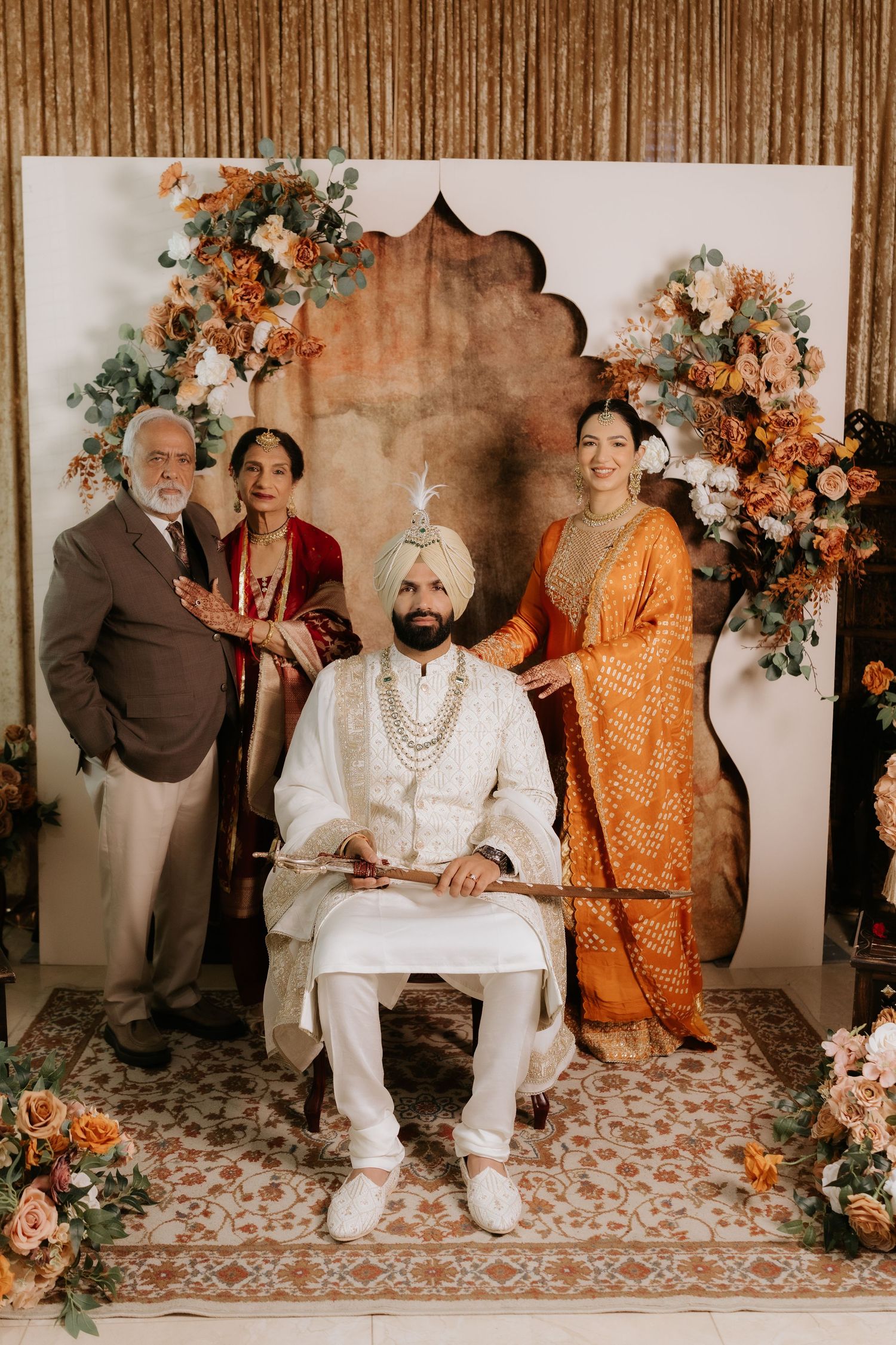 Sikh groom in white sherwani and turban seated with family at traditional Indian wedding ceremony.