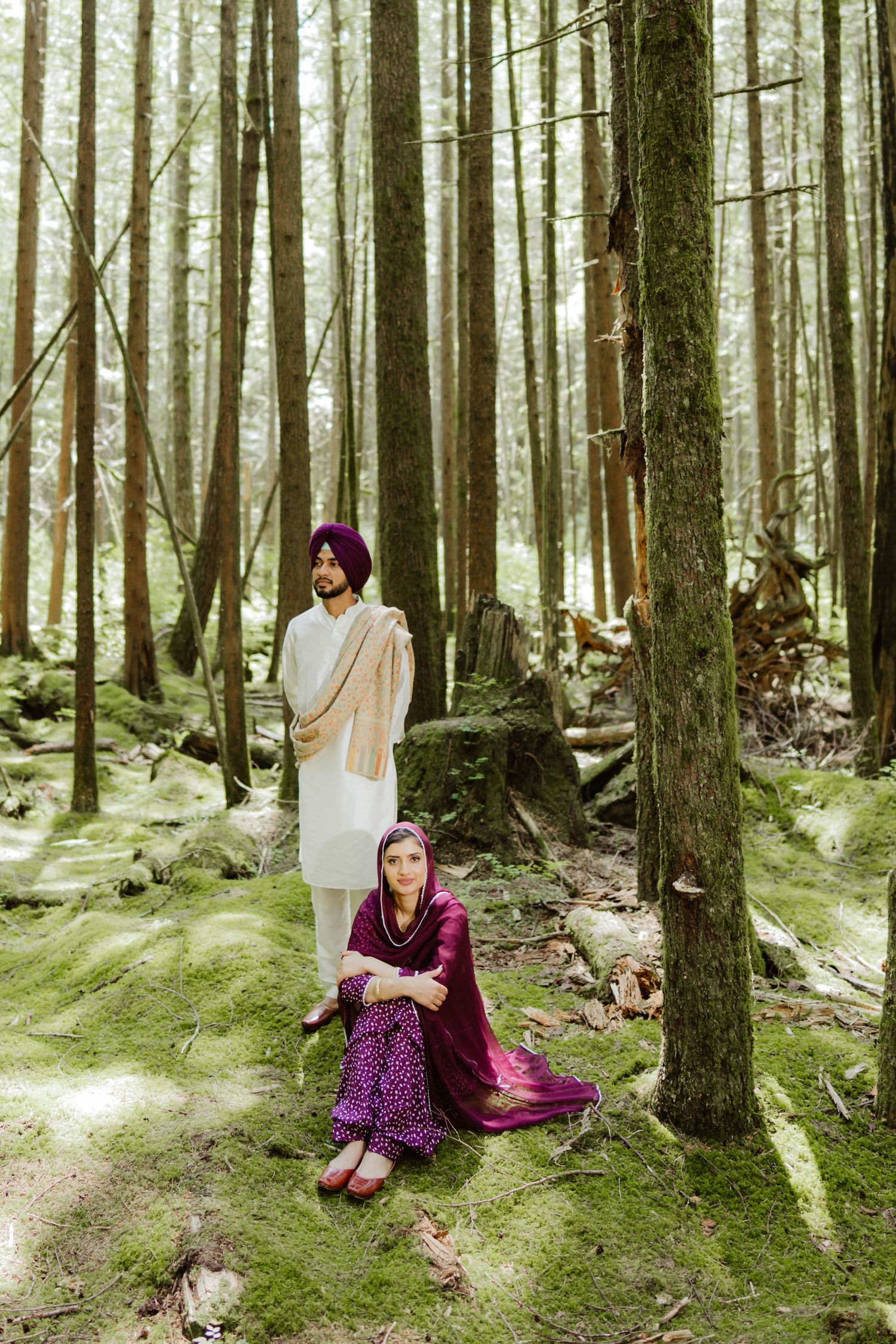 Sikh couple posing in a mossy forest, man standing in white kurta and purple turban, woman seated beside him.