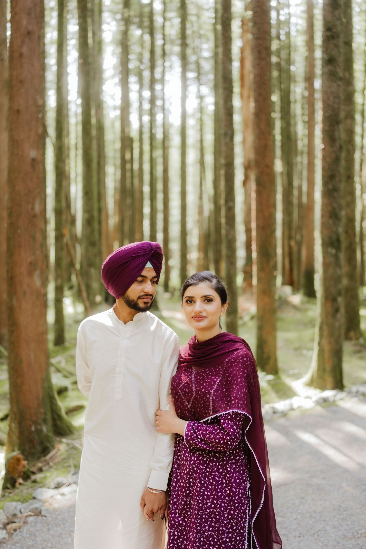 Sikh couple in traditional purple and white outfits posing together on a forest path.