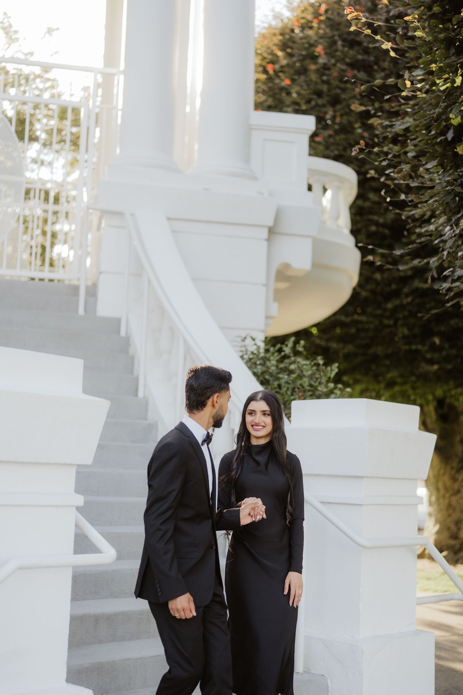 Stylish couple in matching black formal outfits standing on white outdoor staircase surrounded by lush greenery.