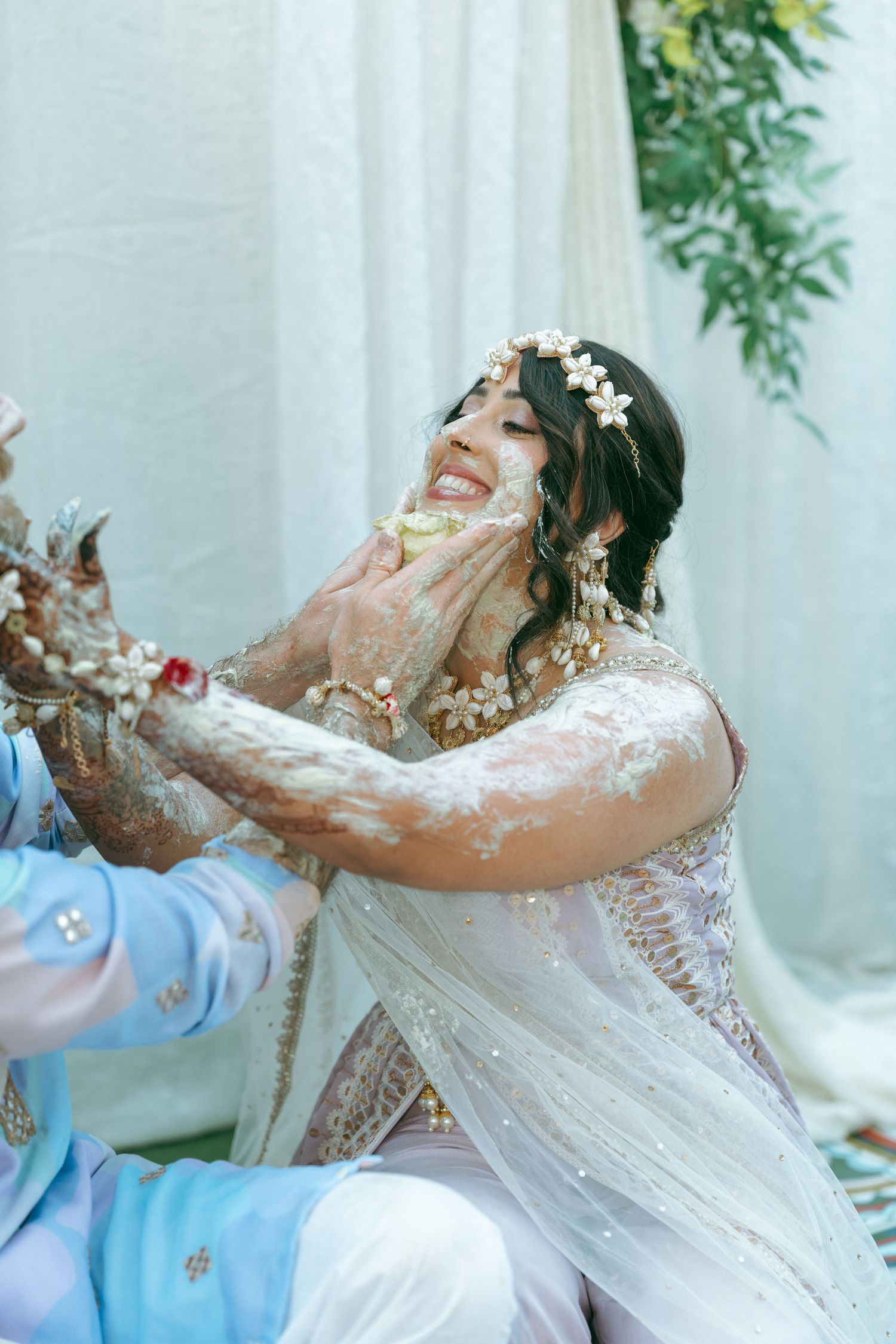 Bride laughing joyfully during Haldi ceremony, covered in turmeric paste, wearing white bridal outfit and floral hair accessories.