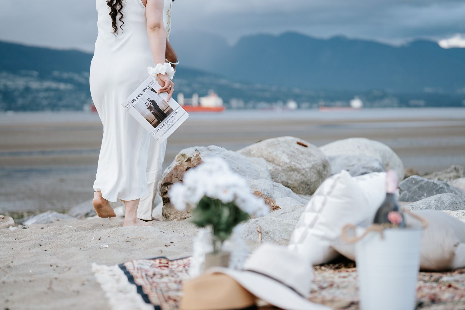 Bride in white dress holds polaroid photos on sandy beach with mountain backdrop and boho picnic setup.