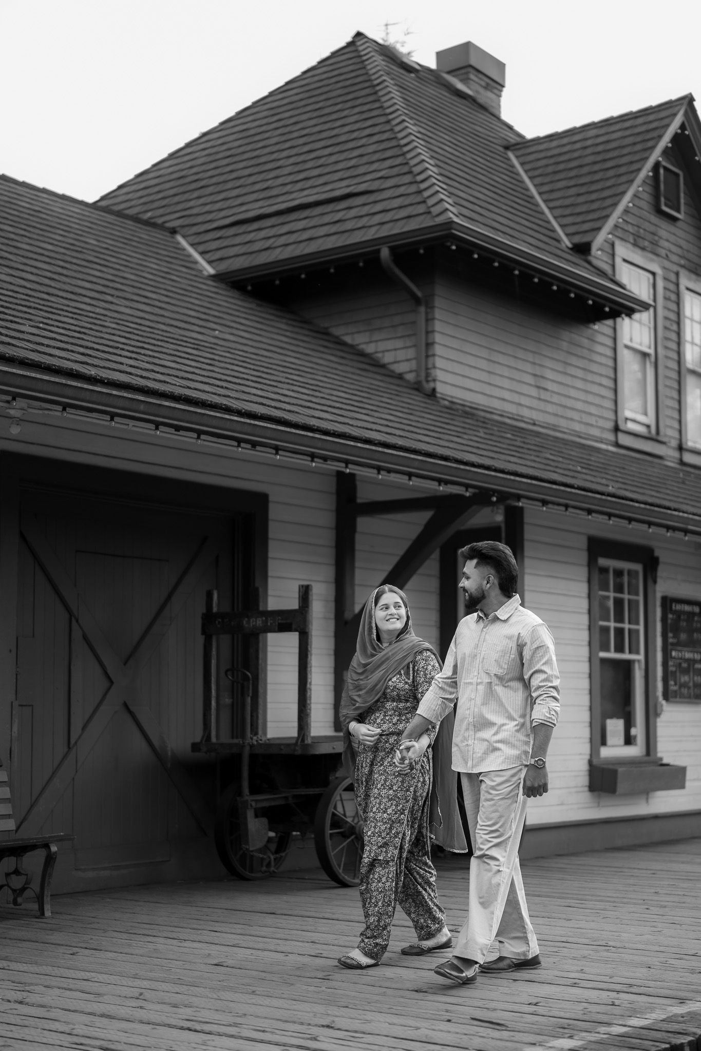 Couple in traditional South Asian attire walking outside historic wooden building, black and white.
