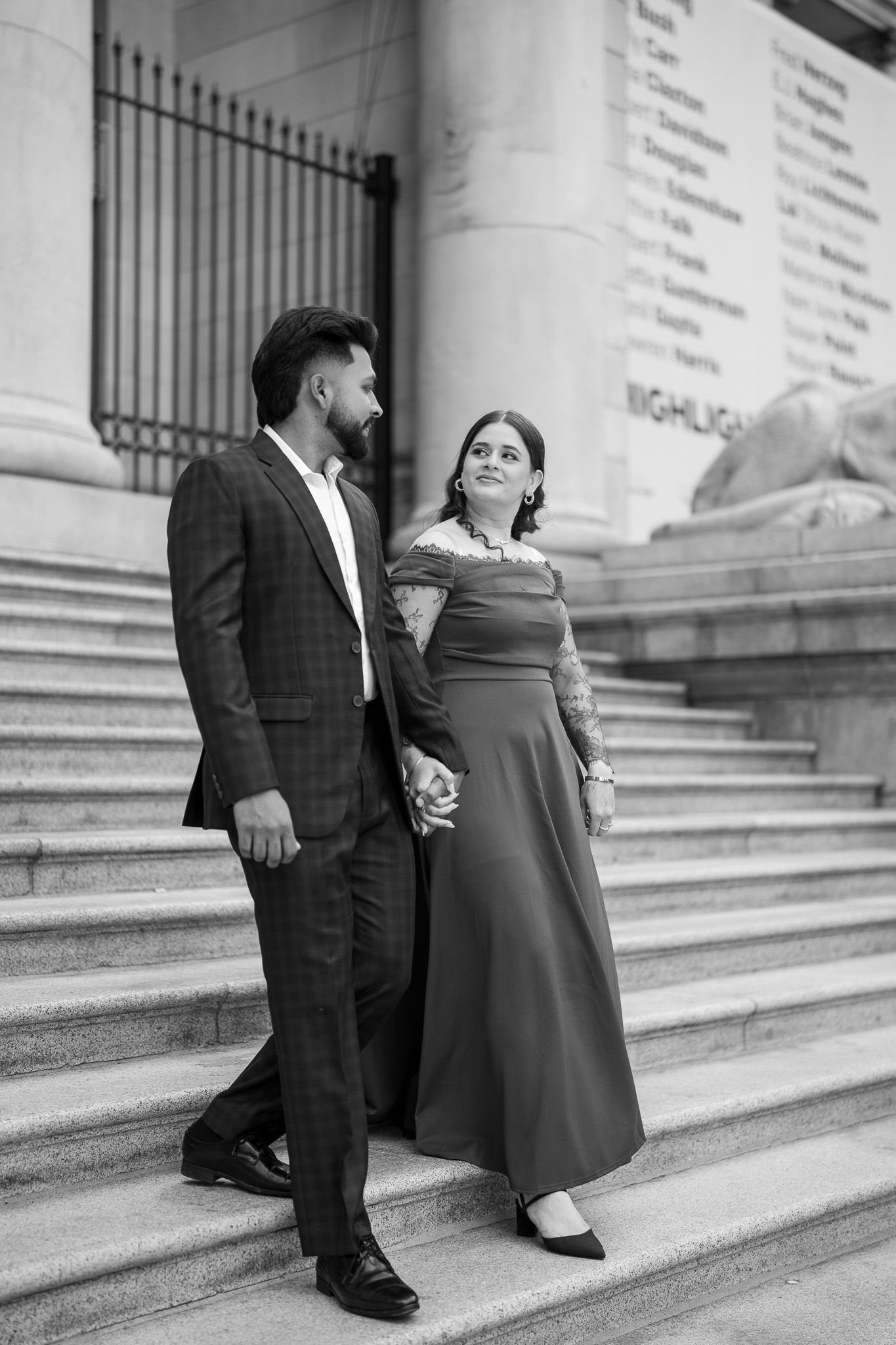 Elegant couple holding hands on steps in black and white, man in plaid suit, woman in long gown.
