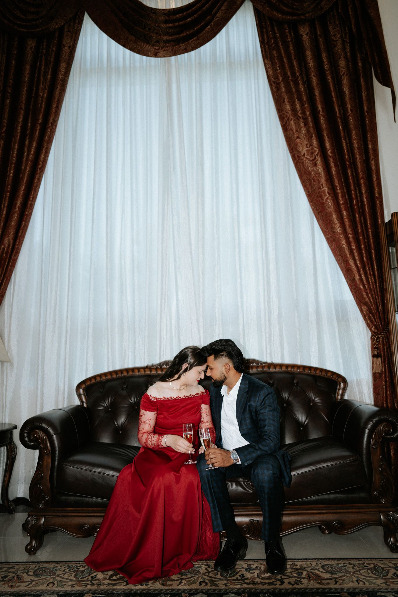 Couple in formal attire sharing champagne on leather sofa with ornate curtains backdrop.