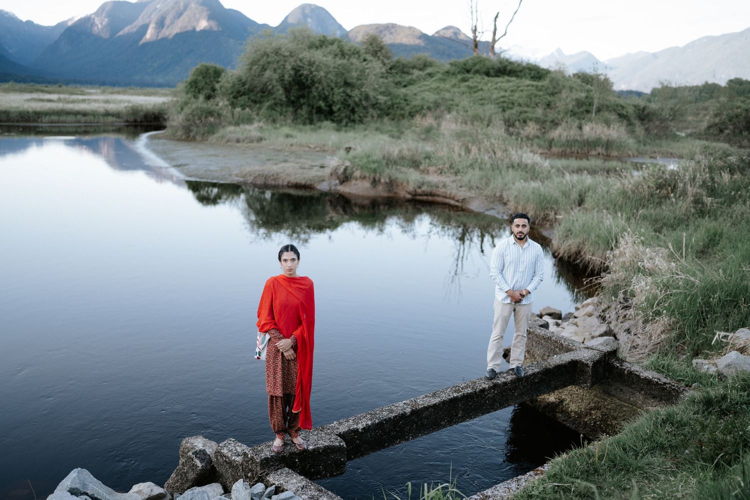Two people stand on a narrow footbridge over calm water with mountains in the background.