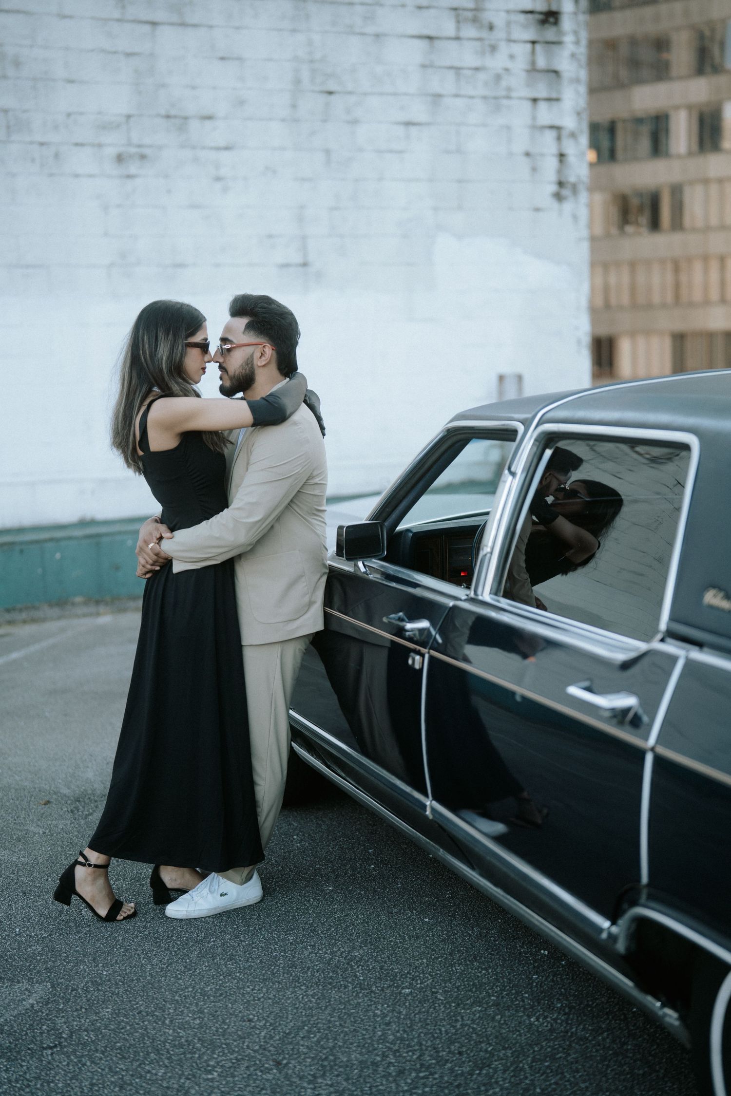 Romantic couple in black and beige outfits pose intimately by vintage black car on urban street.