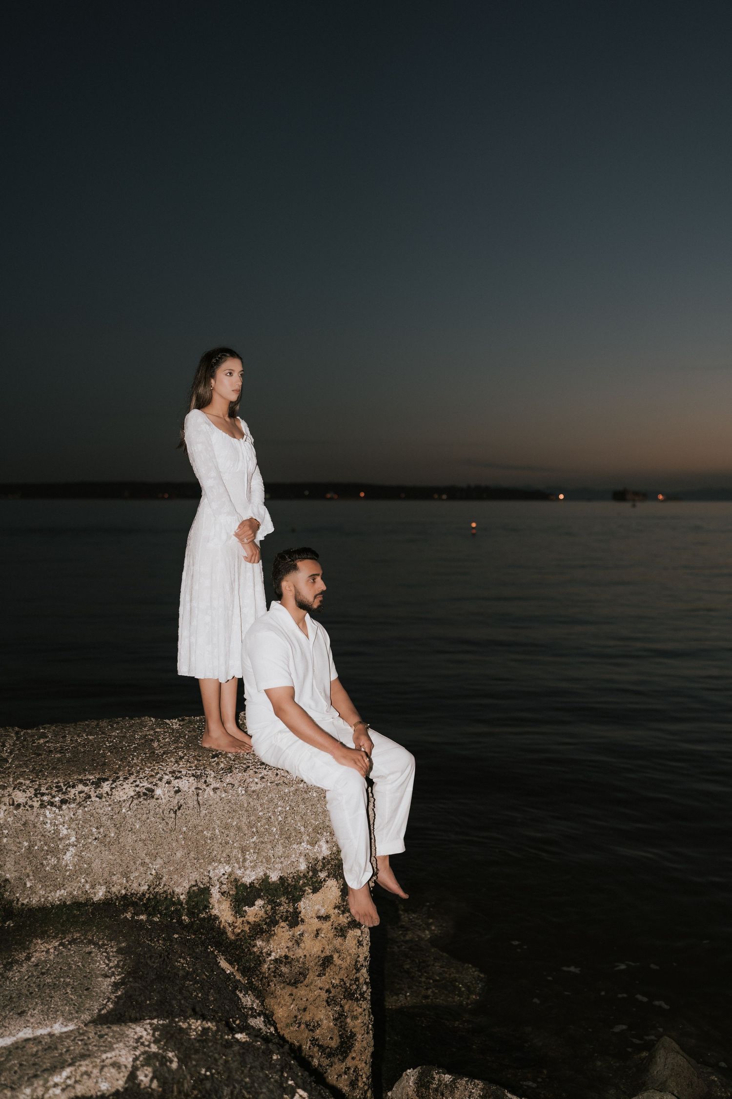 A couple in white outfits sits on coastal rocks at dusk overlooking a calm dark ocean.