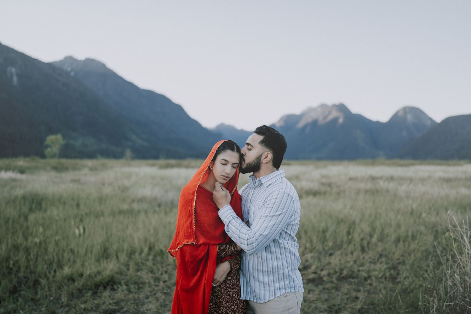 A couple embraces in a grassy meadow with majestic mountain peaks rising in the background at dusk.