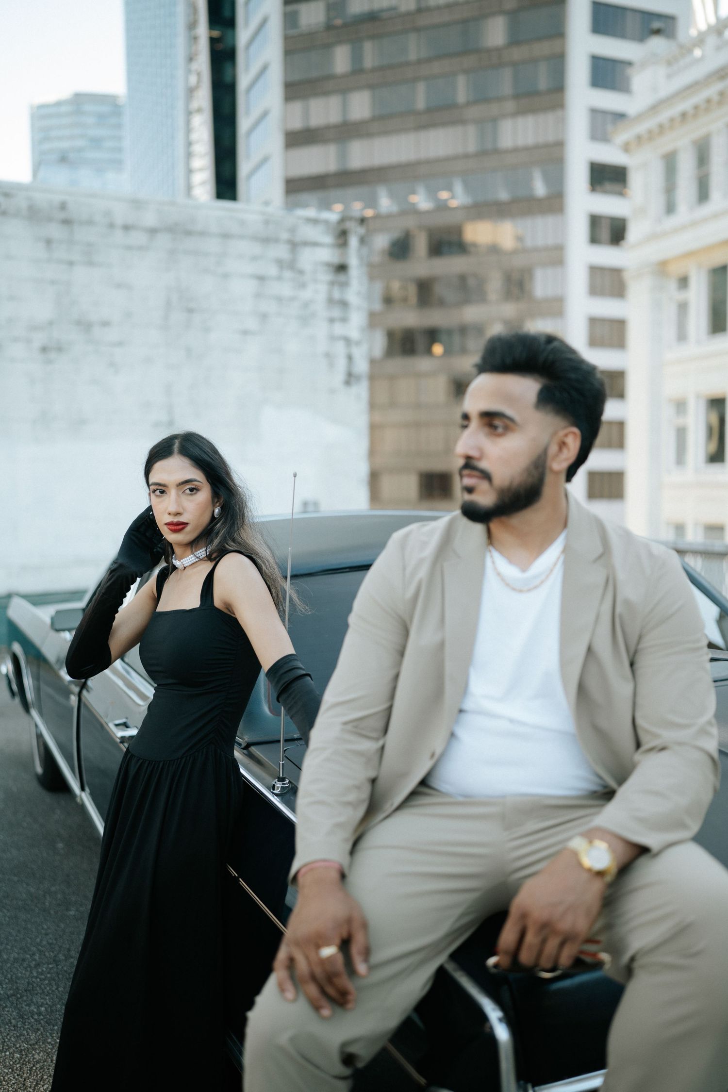 A couple in contrasting black and beige outfits pose together in an urban setting with modern buildings in background.