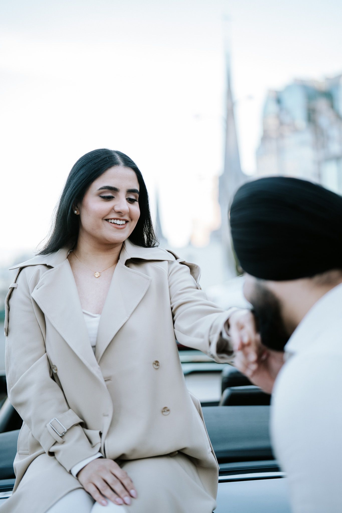 A person in a beige trench coat smiles while sitting outdoors on a sunny day.
