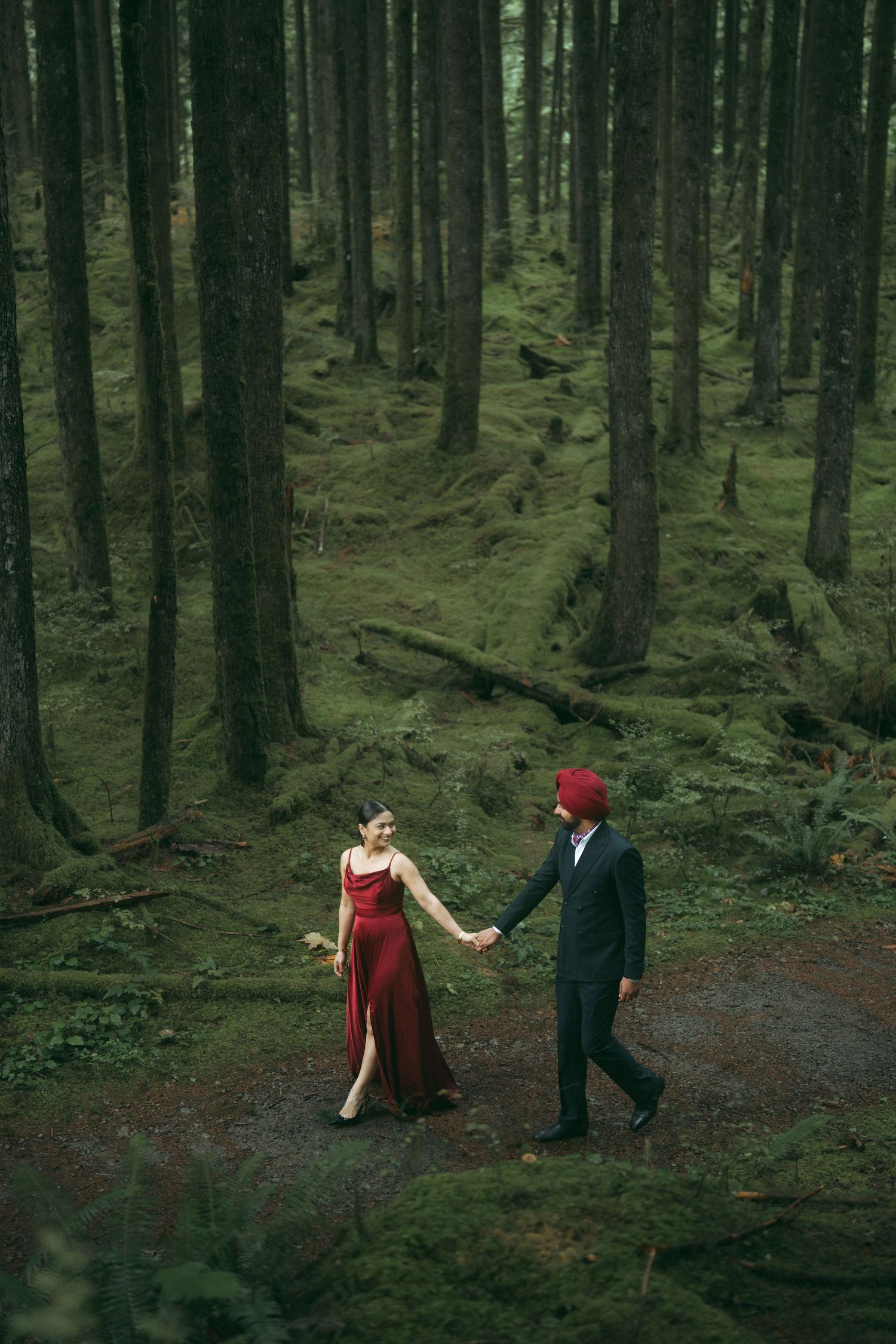 Two figures walk through a mossy forest with tall evergreen trees in dramatic lighting.
