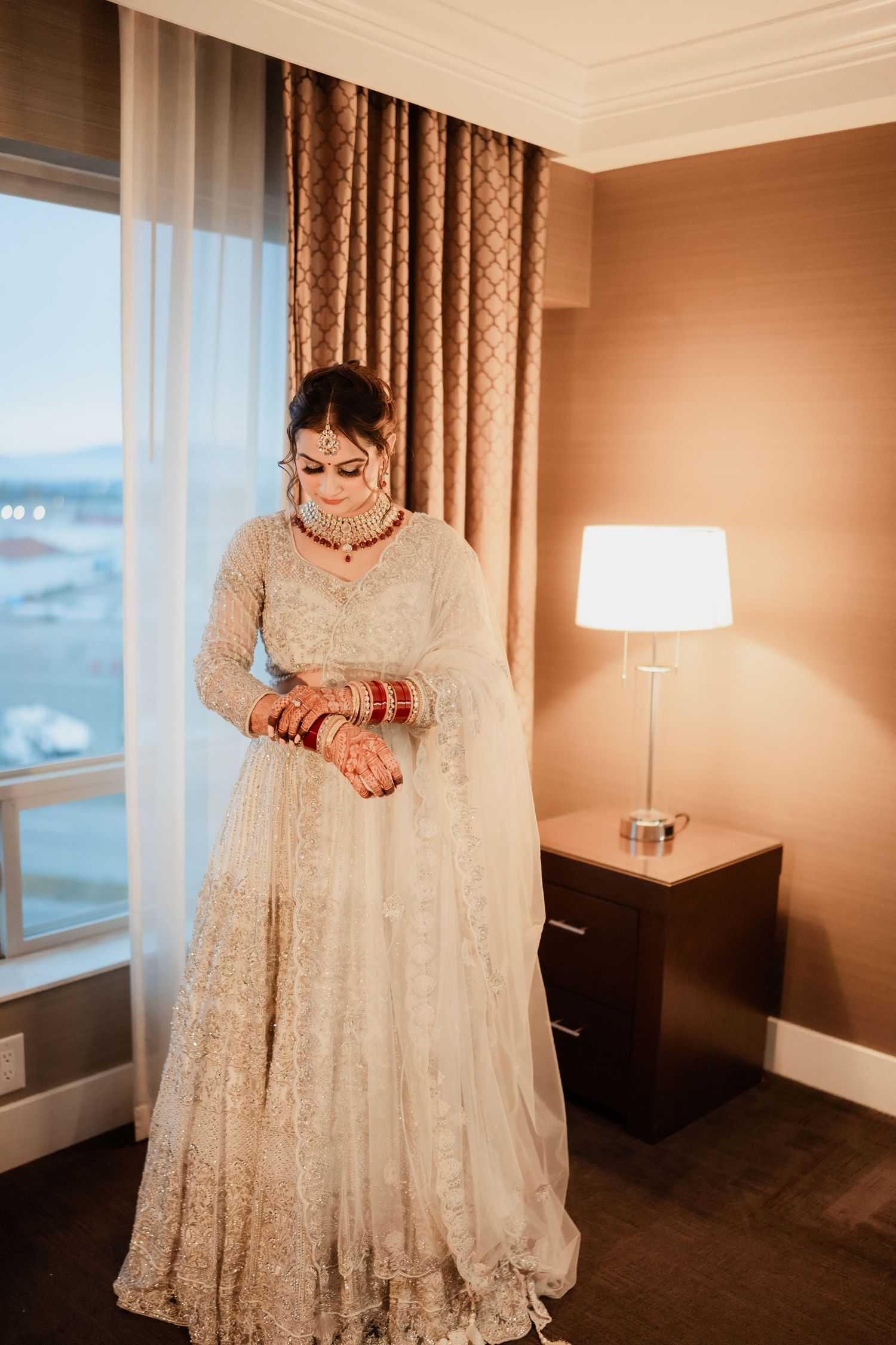 A bride in a detailed white lehenga showcases her traditional bridal bangles in a sunlit room.