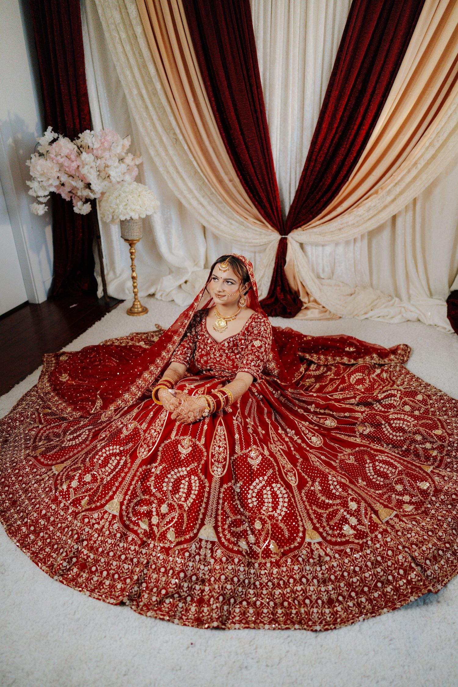 Luxurious red and gold bridal lehenga displayed against cream and burgundy draped backdrop.