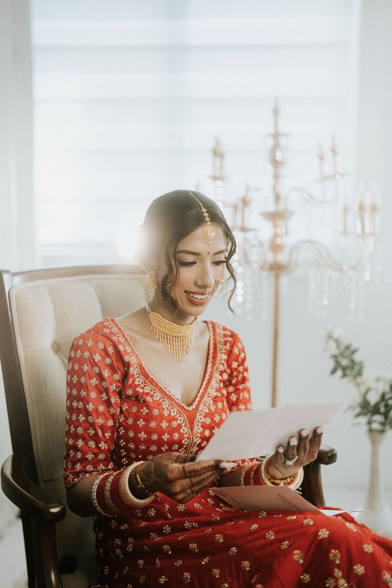 Traditional Indian bride in red lehenga reads a letter while sitting in an elegant white room with candelabras.
