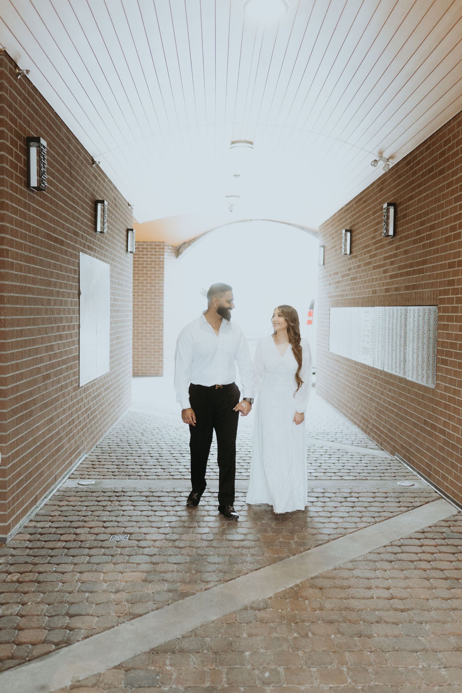 A couple walks down a brick-lined hallway in elegant white and black attire, creating a romantic atmosphere.