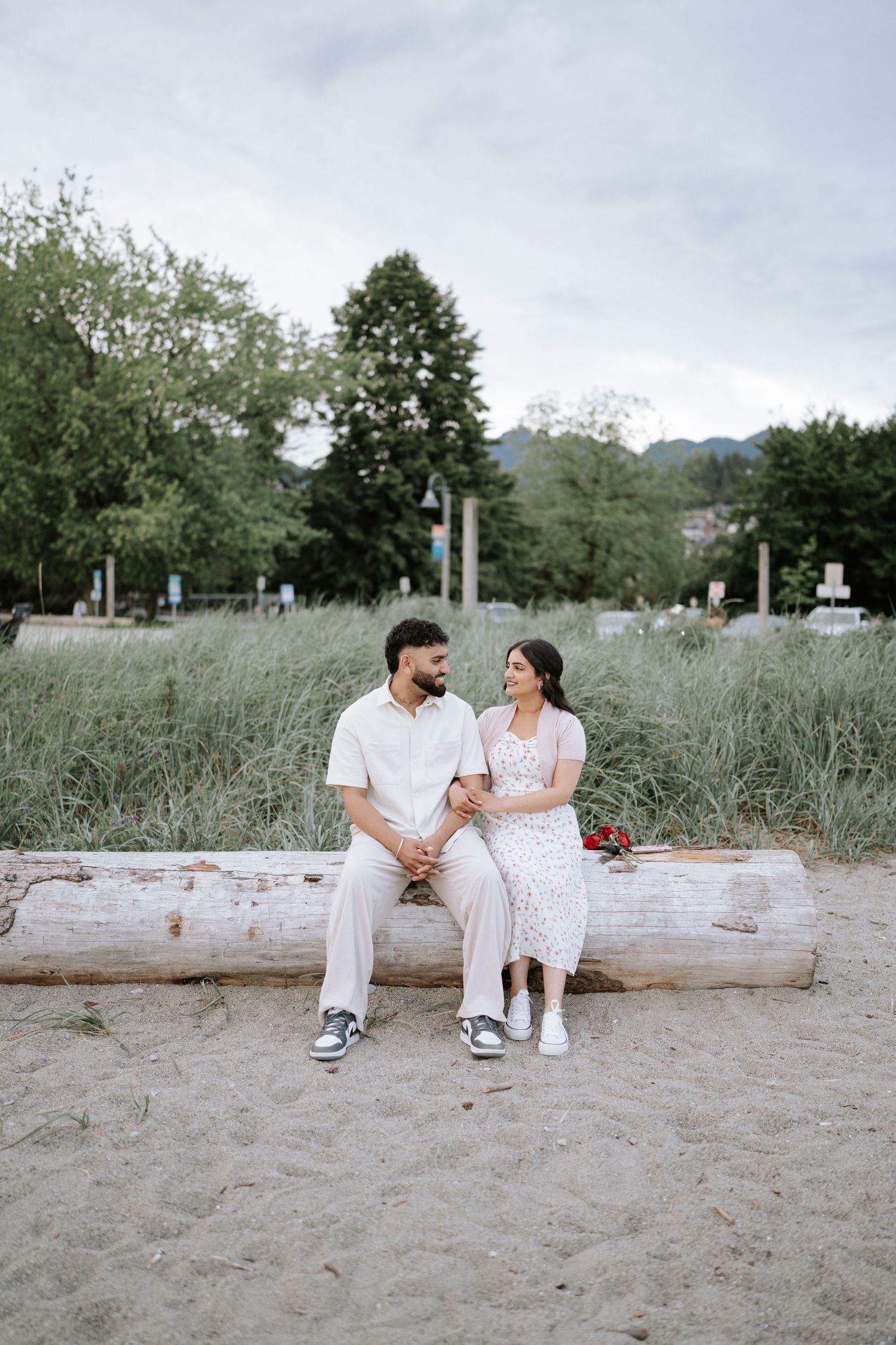Two people in white outfits share intimate moments on a beach log surrounded by grass.