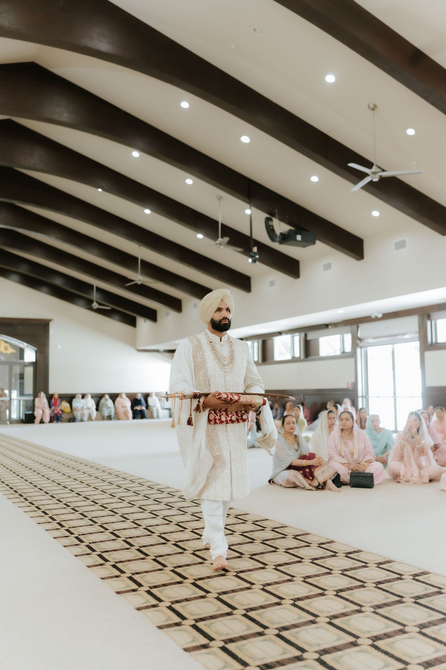 Sikh groom in white sherwani and turban walks down the aisle during a traditional Anand Karaj wedding ceremony.