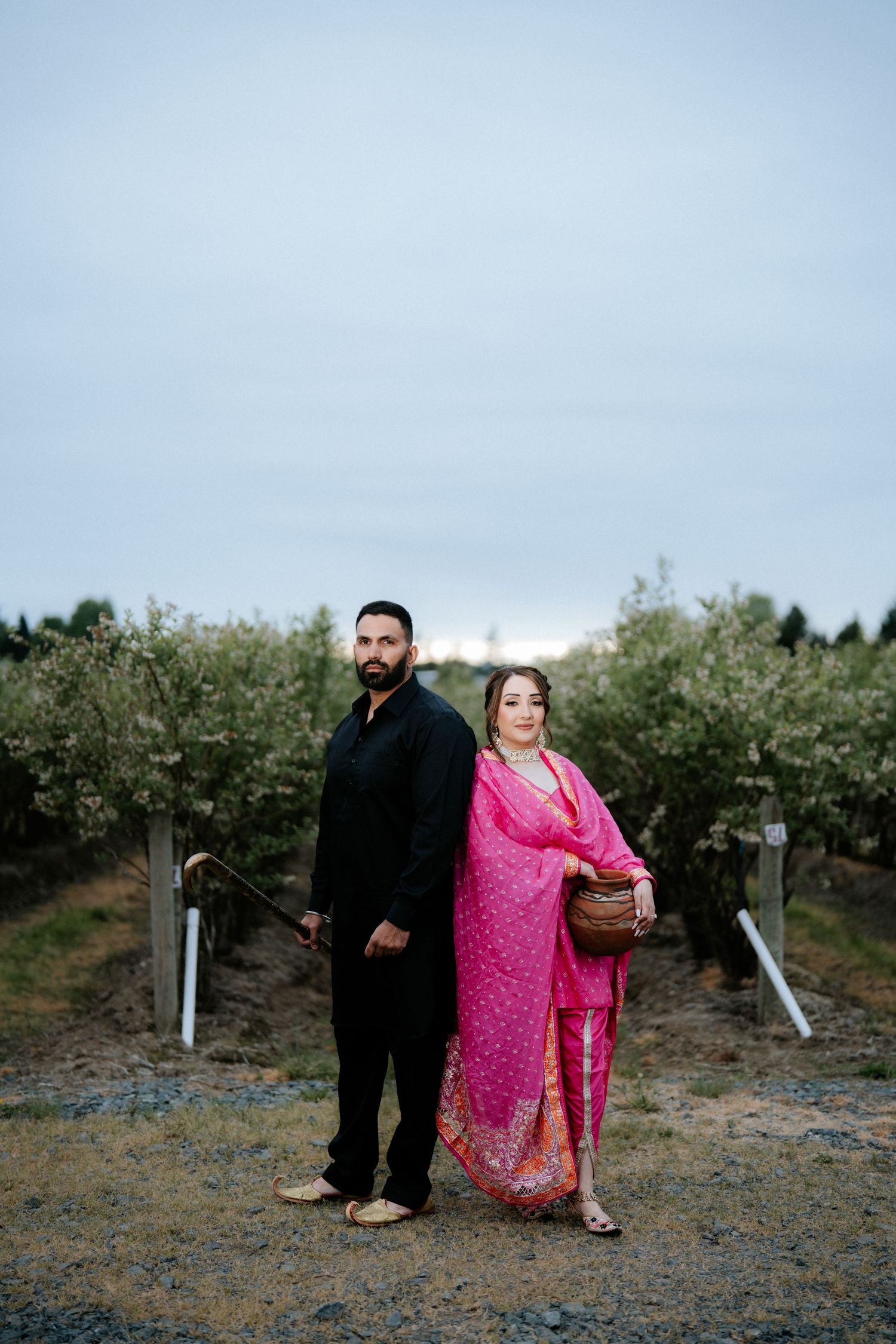 Couple in traditional attire posing in an orchard, man in black and woman in pink saree, at dusk.