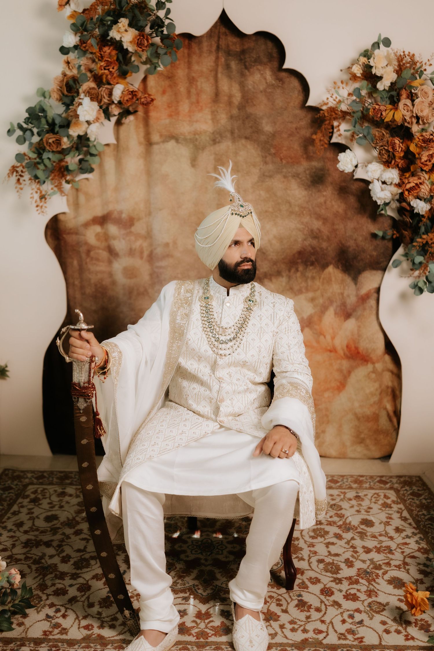 Sikh groom in white sherwani and turban sits regally on ornate chair with floral backdrop at traditional wedding.