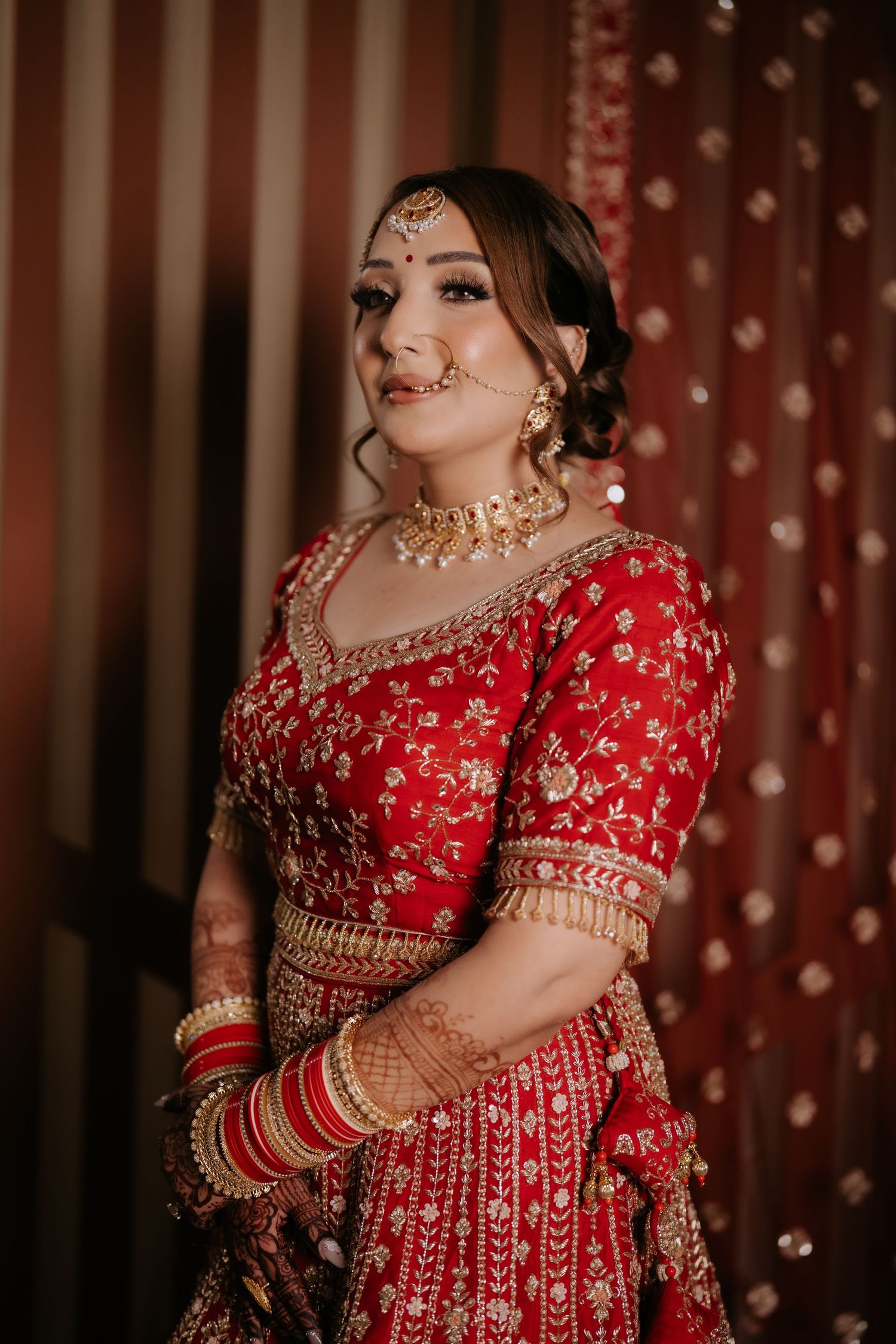 A bride in a red embroidered lehenga with gold jewelry, maang tikka, and mehndi poses gracefully at her wedding.