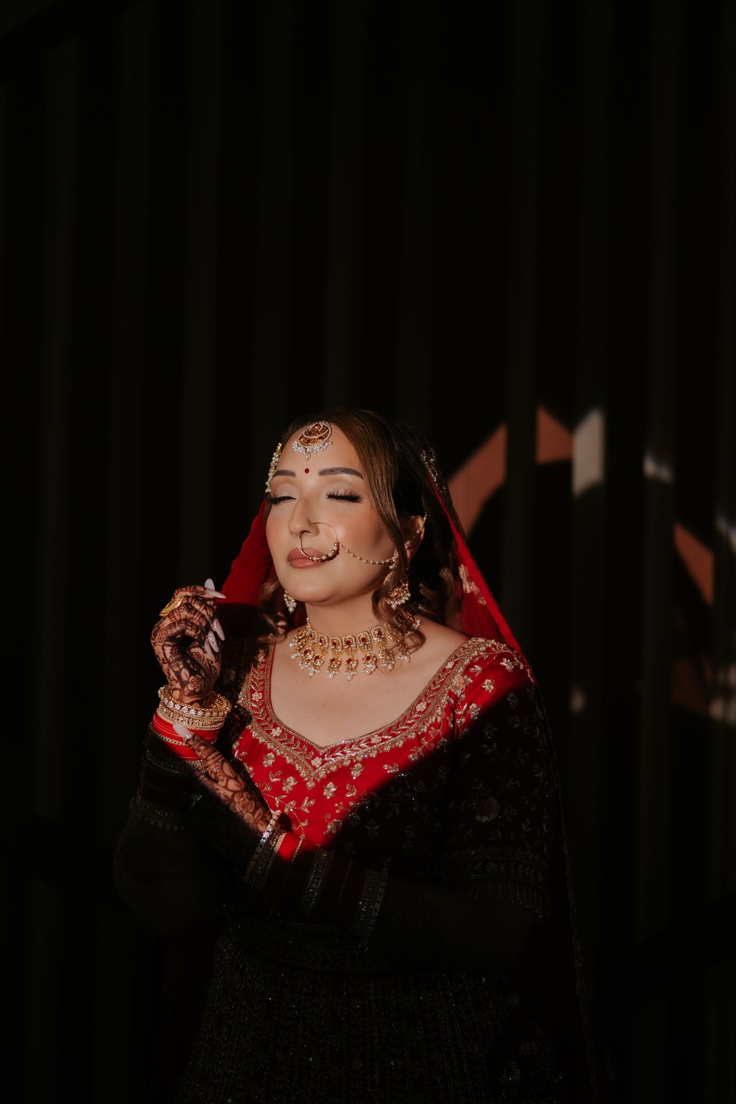Indian bride in red and black lehenga with gold jewelry and maang tikka poses elegantly against dark backdrop.