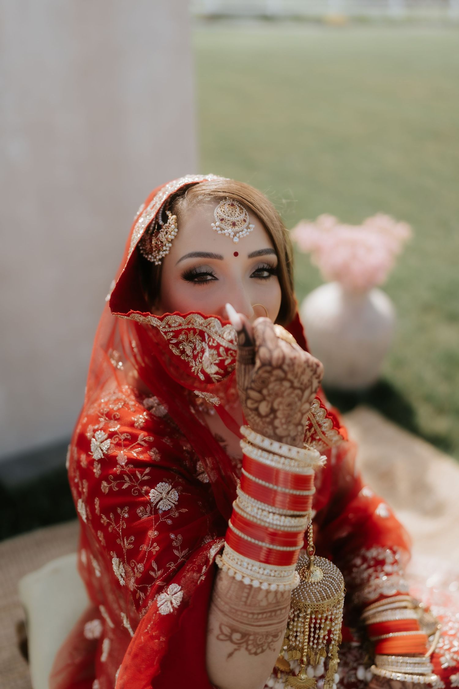 Indian bride in red bridal lehenga with gold embroidery, wearing bangles and maang tikka, posing with mehndi-adorned hands.