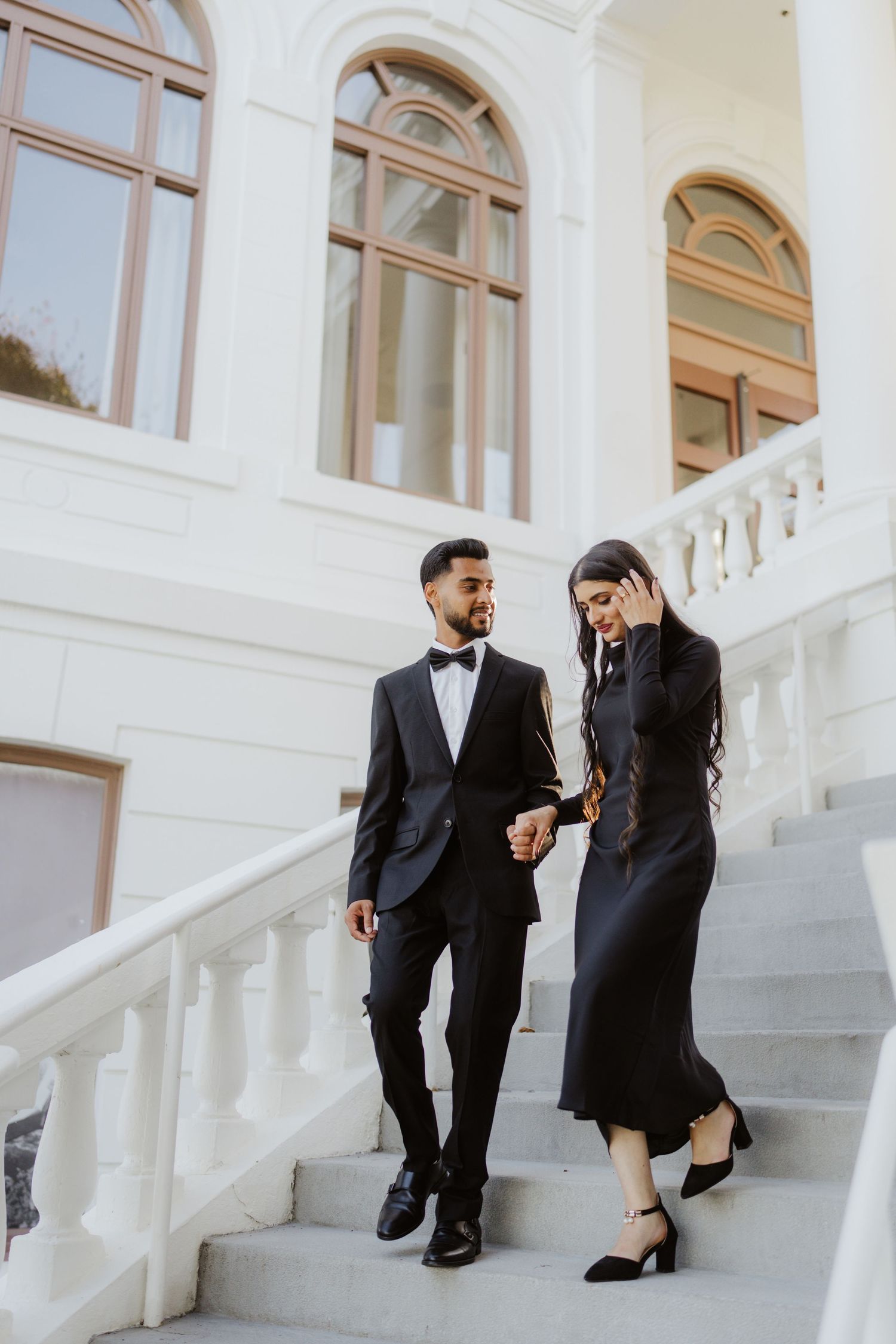 Elegant couple in matching black formal attire walking down white marble stairs of a classic building.