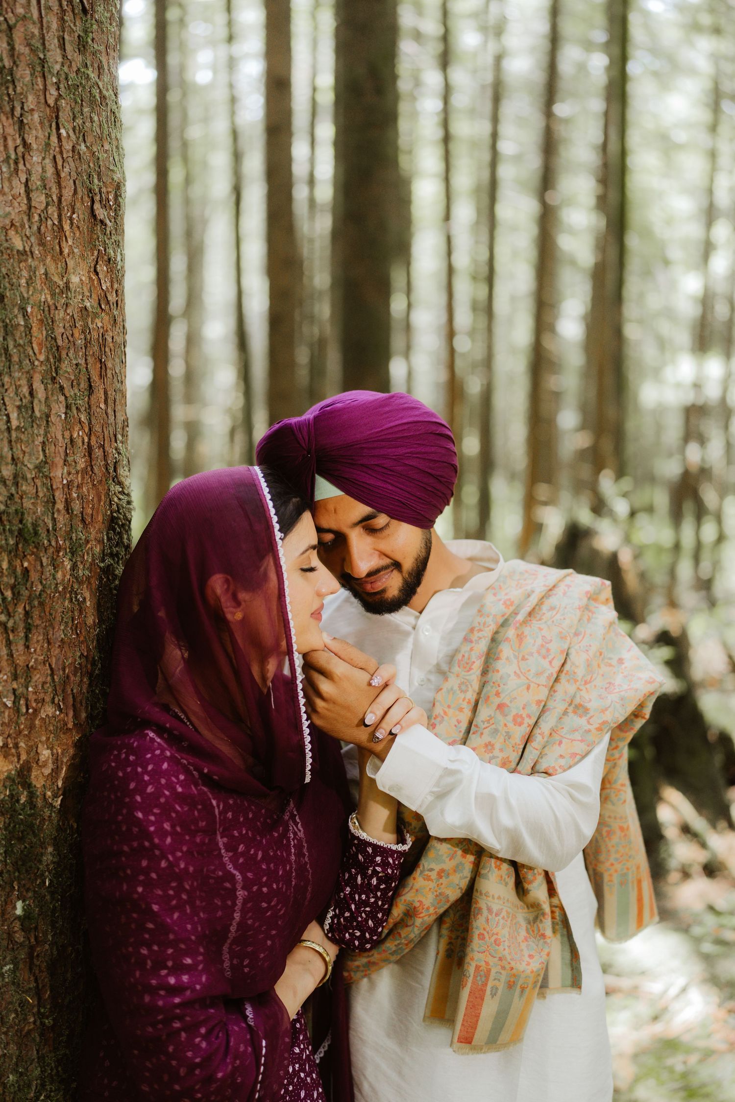 Sikh couple in traditional attire sharing tender moment leaning against tree in forest.