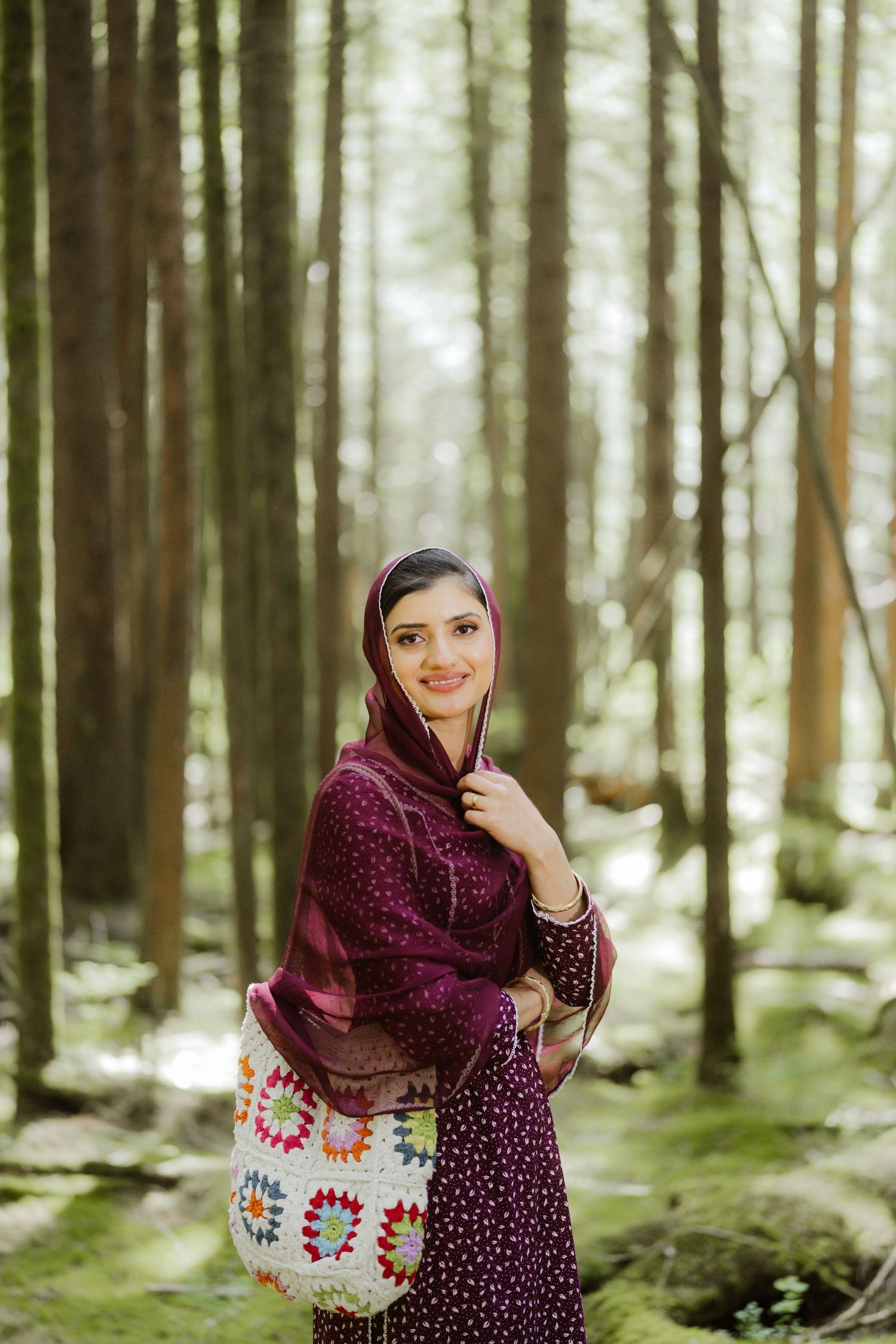 Smiling South Asian woman in purple traditional dress holding a colorful tote bag in a forest.