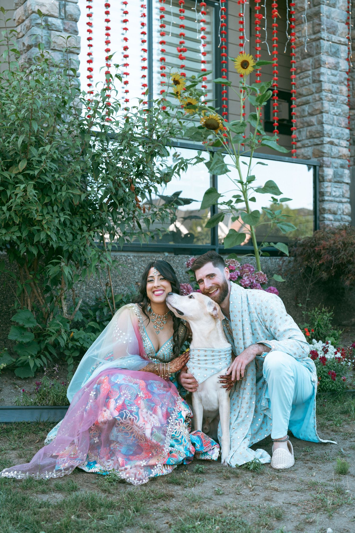 Couple in traditional Indian attire sitting outdoors with a white dog, surrounded by floral decorations and sunflowers.