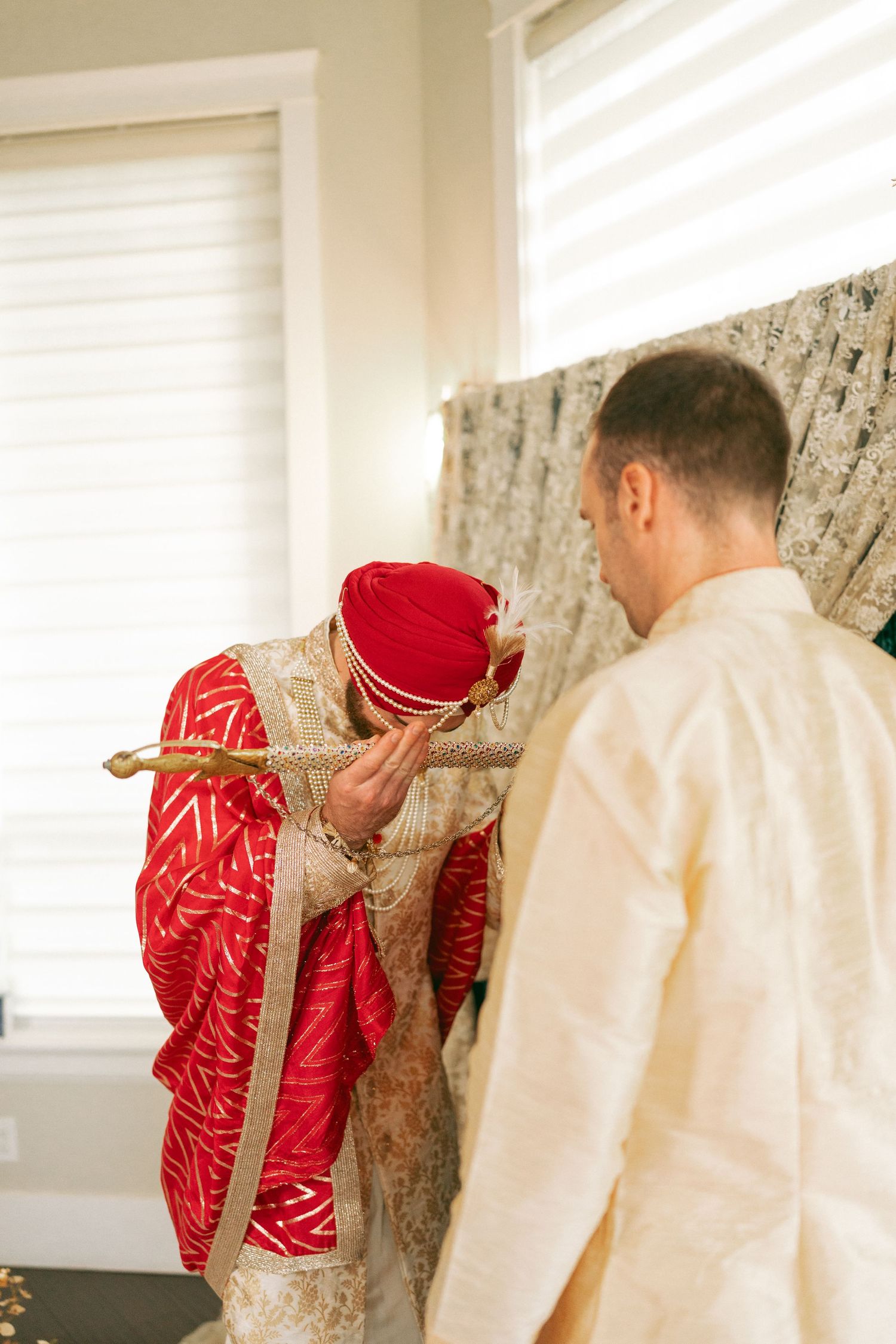 Groom in red sherwani and turban bowing respectfully before a man in cream kurta during an Indian wedding ceremony.