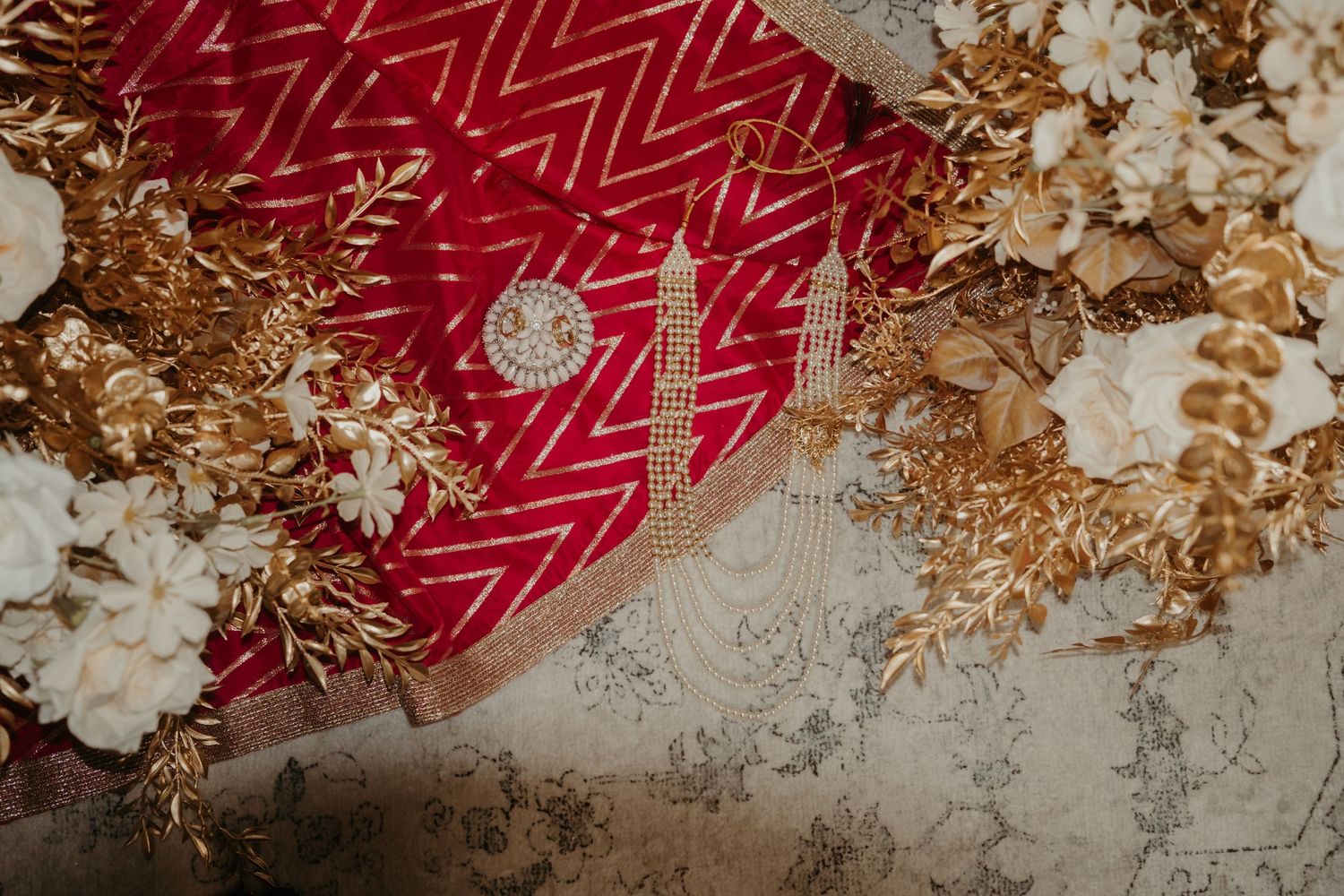 Red velvet bridal lehenga with gold embroidery, surrounded by golden floral accessories and white blooms.