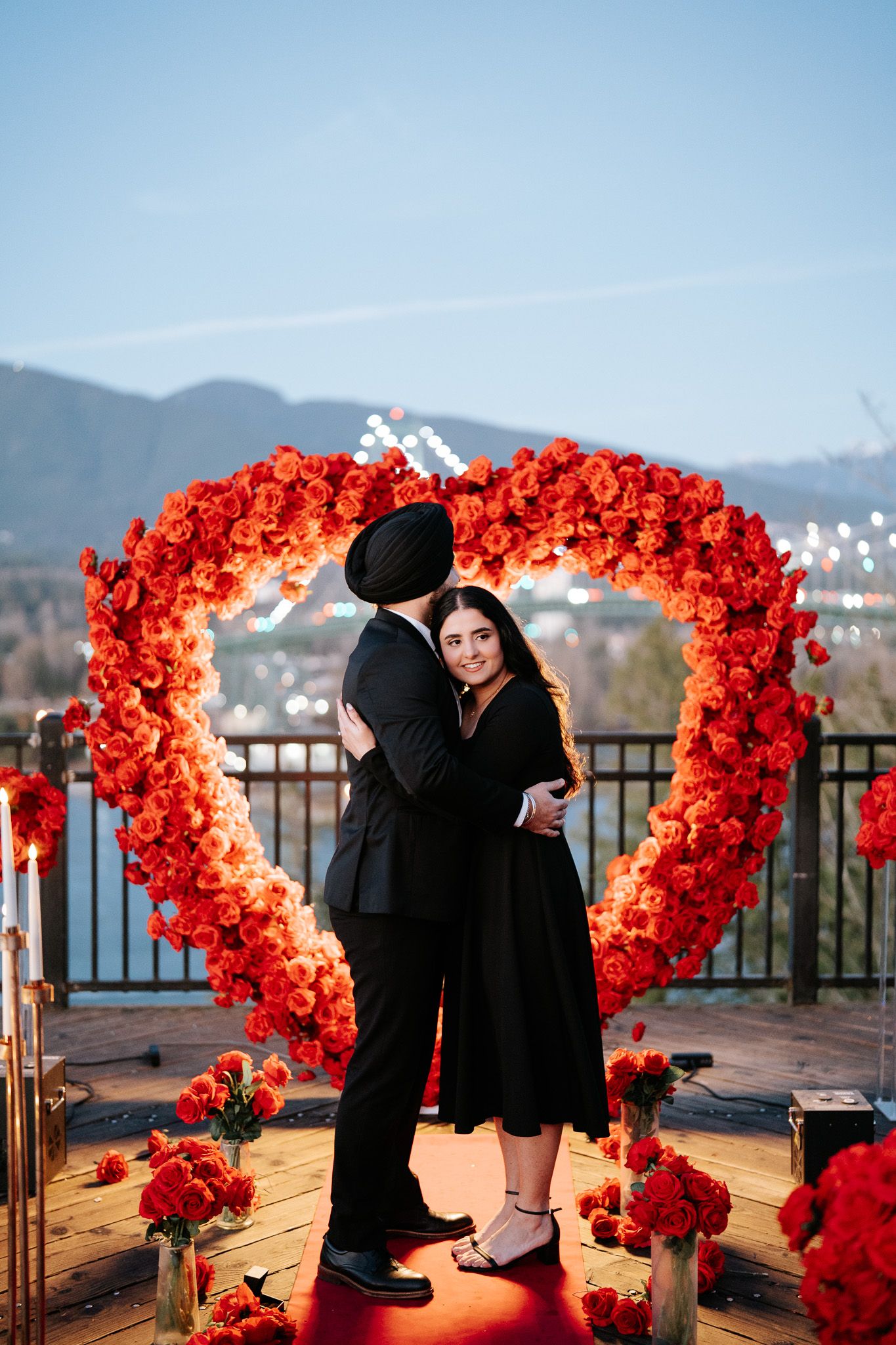 A romantic proposal sequence under vibrant red rose arches on a rooftop with mountain views in the background.