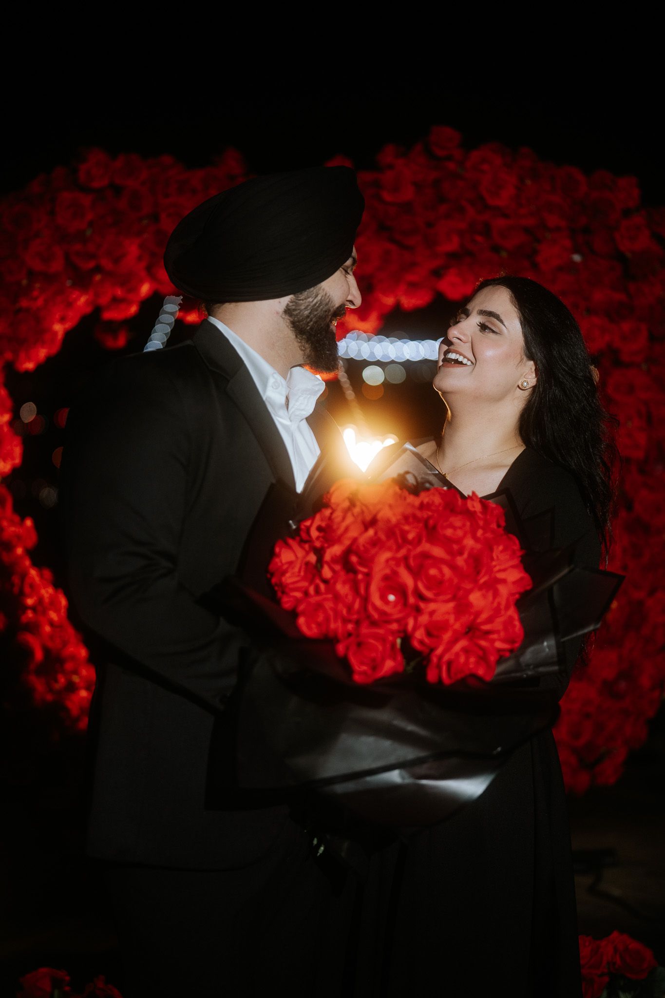 A romantic couple embraces against a backdrop of vibrant red roses in a dramatic silhouetted photoshoot.