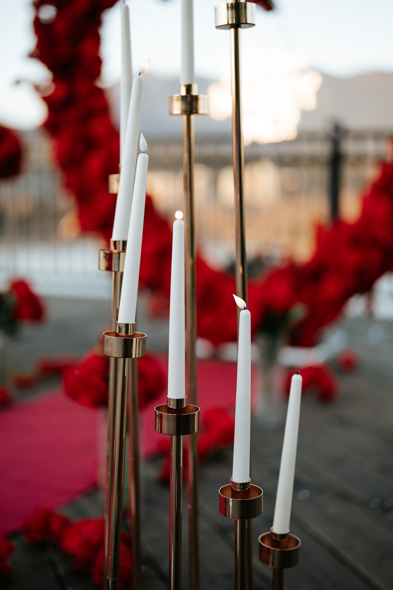 White candles in gold candlesticks stand against a blurred background of red flowers.