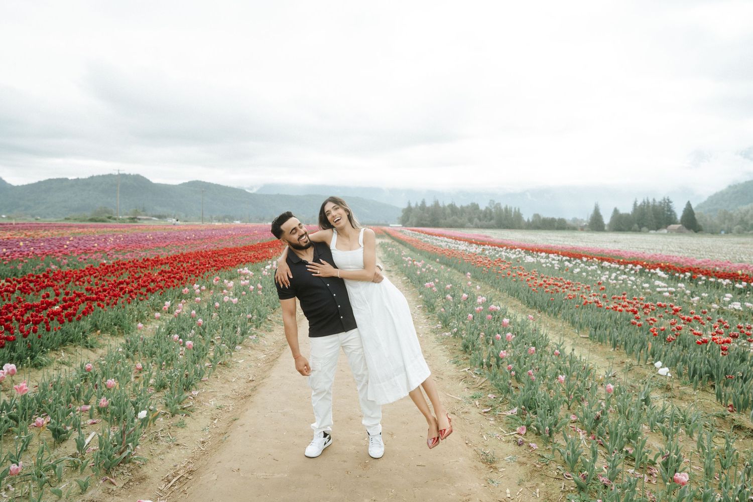 A couple walks hand in hand through vibrant red tulip fields with mountains in the misty background.