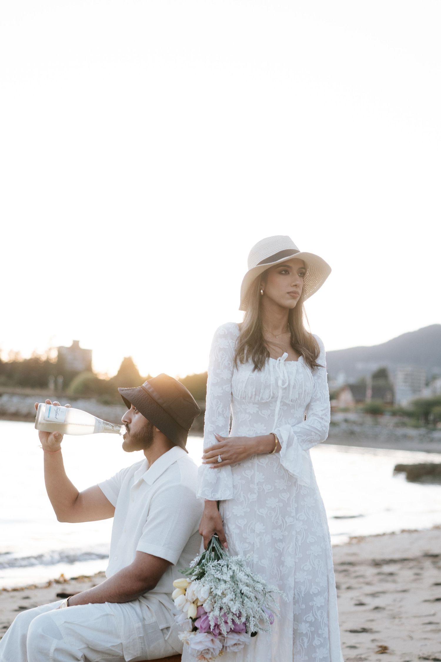 Couple in white outfits having romantic moment on beach at sunset with flowers and mountains in background.