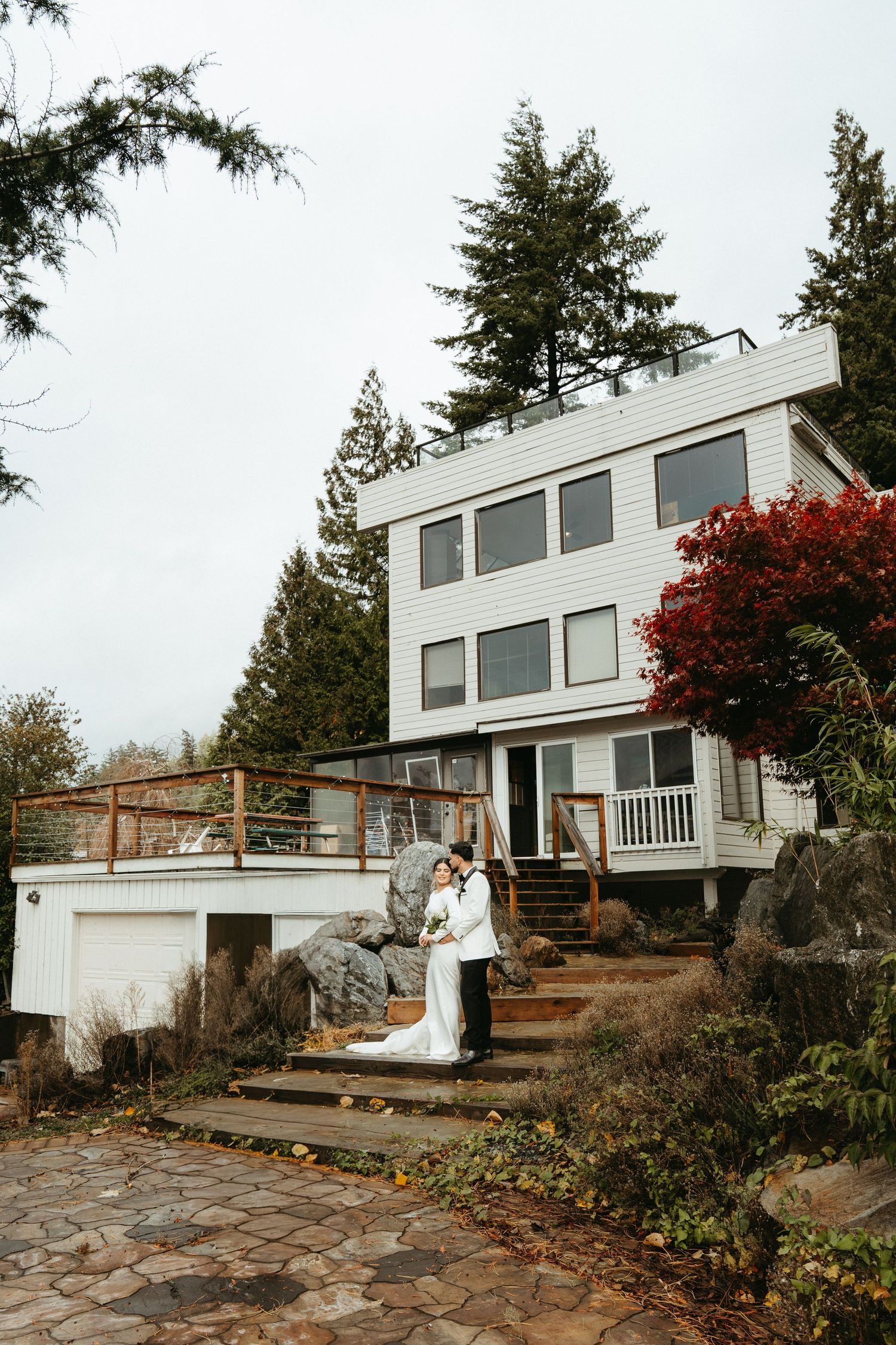 Modern A-frame house with large windows and stone patio surrounded by evergreen trees in Pacific Northwest setting.