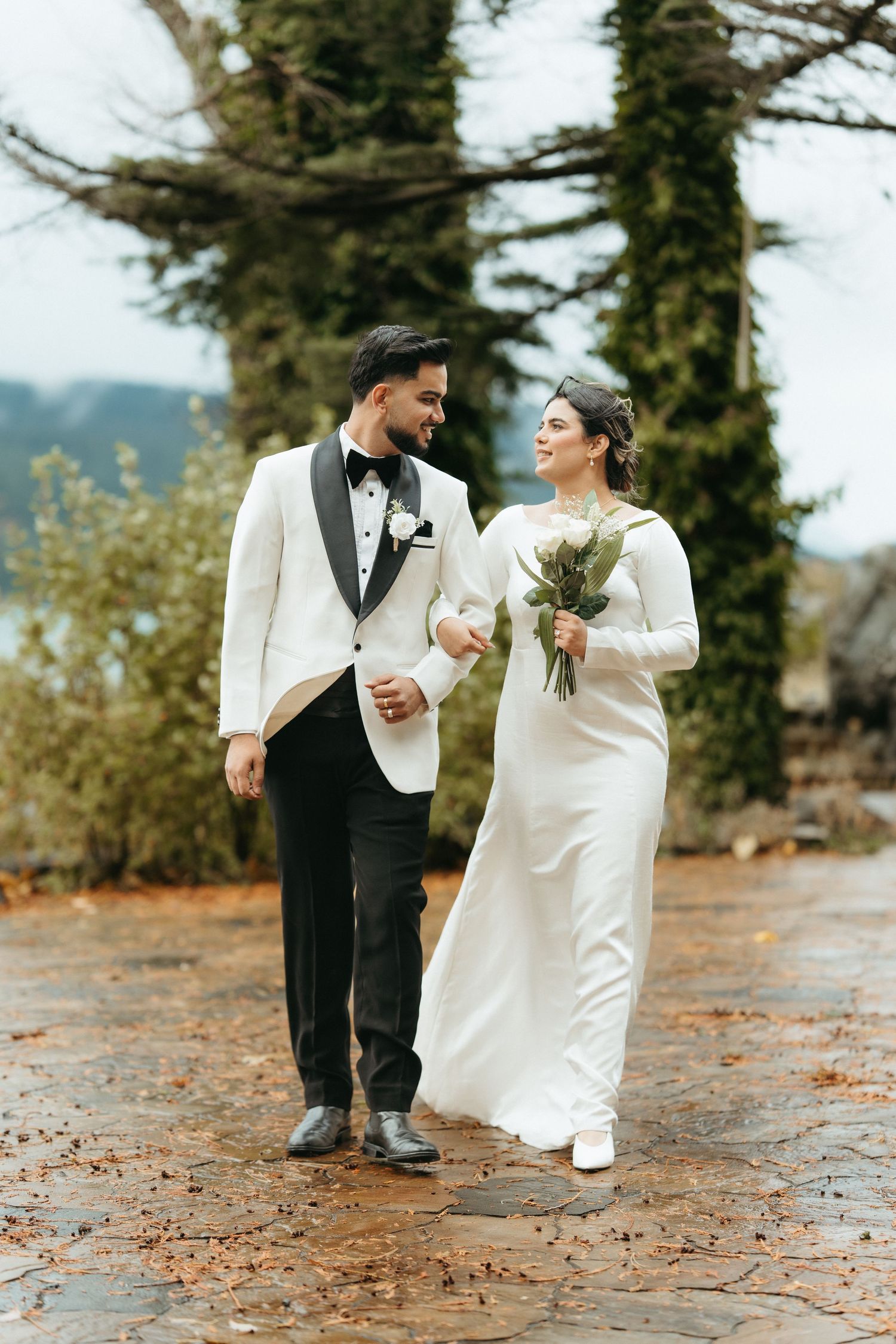 A couple in wedding attire shares a joyful moment together on a leaf-covered path in an outdoor setting.