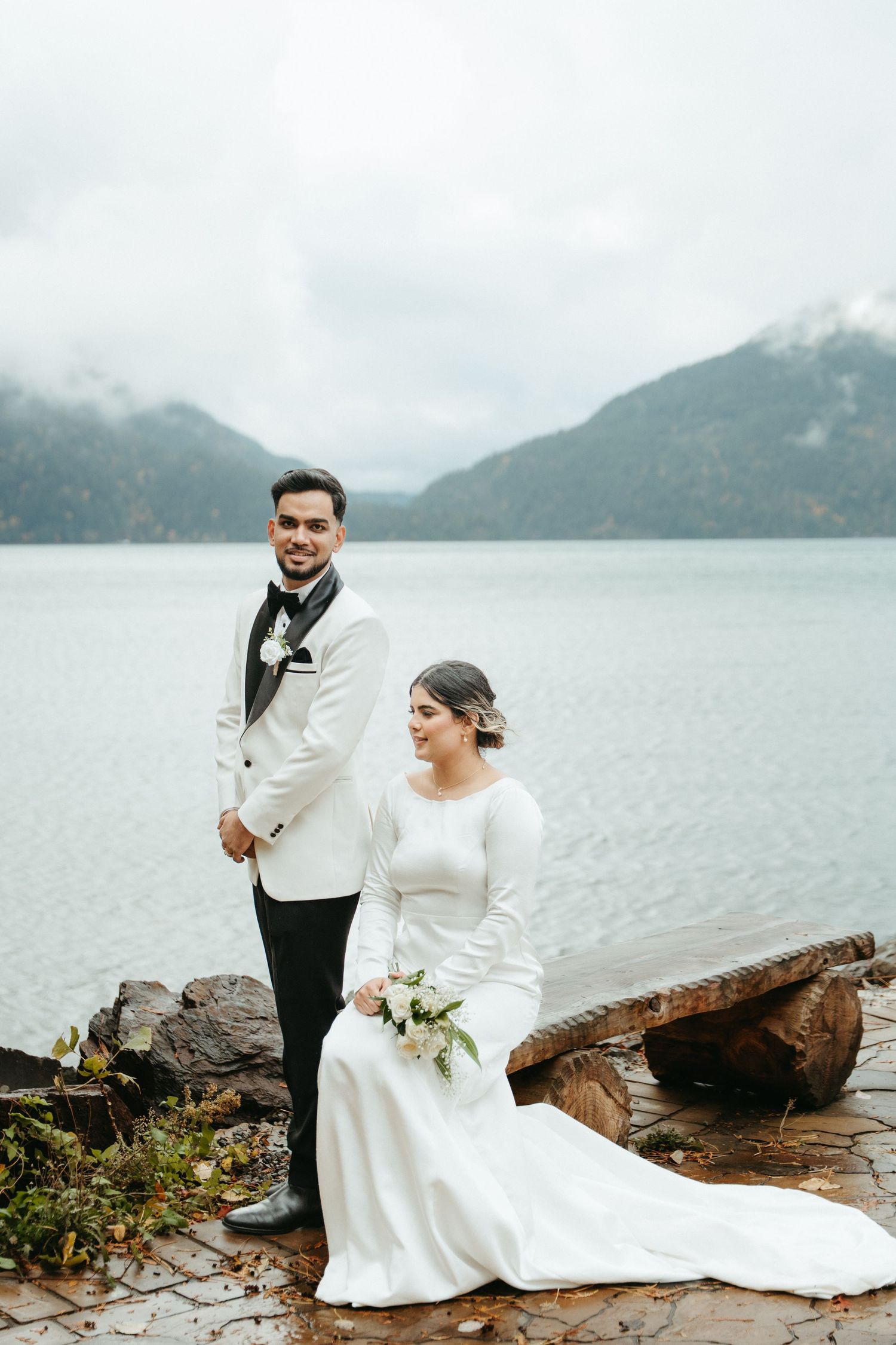 A couple in wedding attire poses on rocky shoreline with misty mountains and lake in the background.
